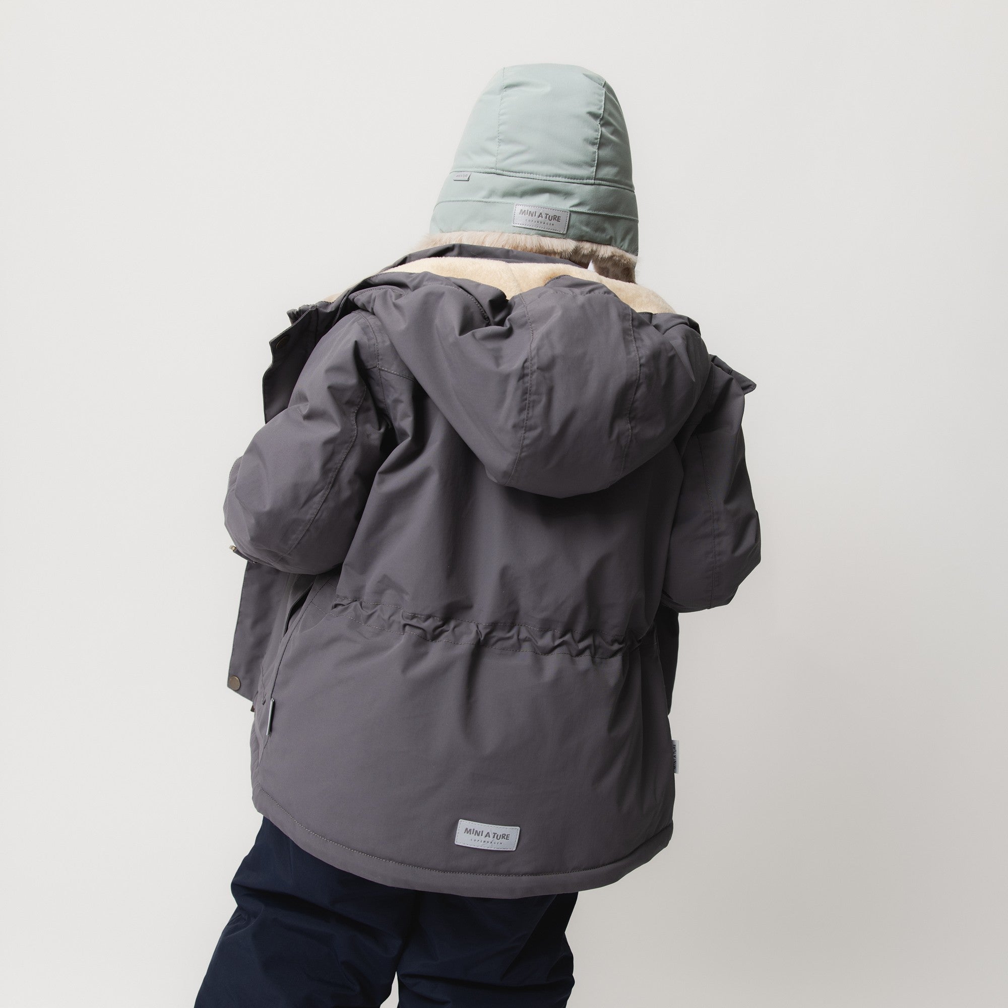MATWALDO fleece lined winter jacket in charcoal grey worn by a child shown from the back, with the detachable hood fastened, beige fleece lining visible at the collar, adjustable internal waist creating a gathered silhouette at the lower back and a white MINI A TURE label at the hem.