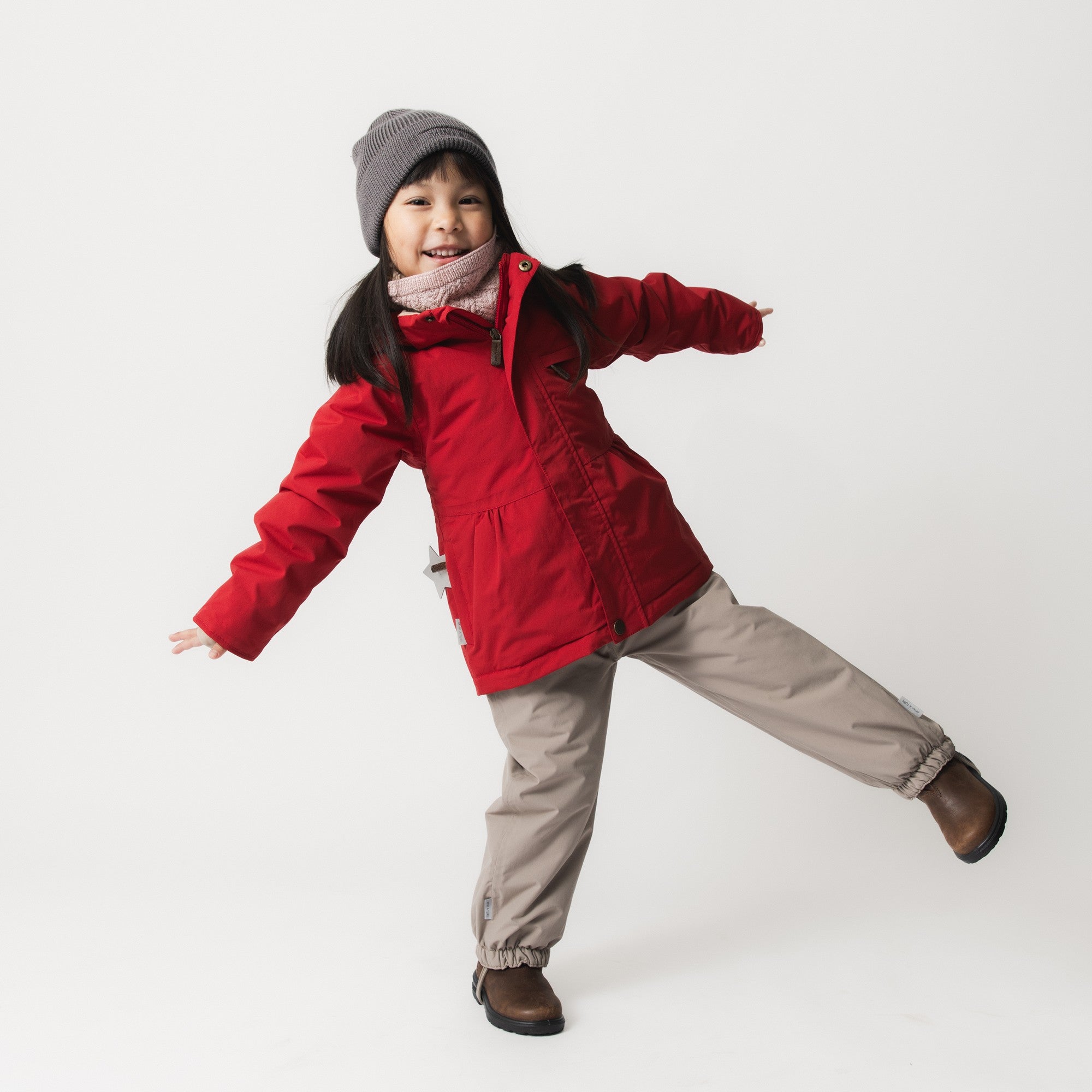 Young girl wearing the MATBRIDDINE winter jacket in red with soft gathers at the waist, paired with beige trousers, brown boots and a grey knitted hat, captured mid-movement in a playful pose.
