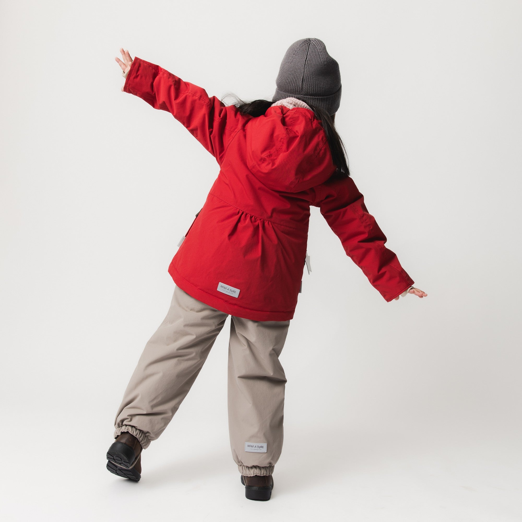 Young girl wearing the MATBRIDDINE winter jacket in red with gathered waist detail, shown from behind with arms outstretched in an energetic pose, paired with beige trousers, black boots and a grey knitted beanie.