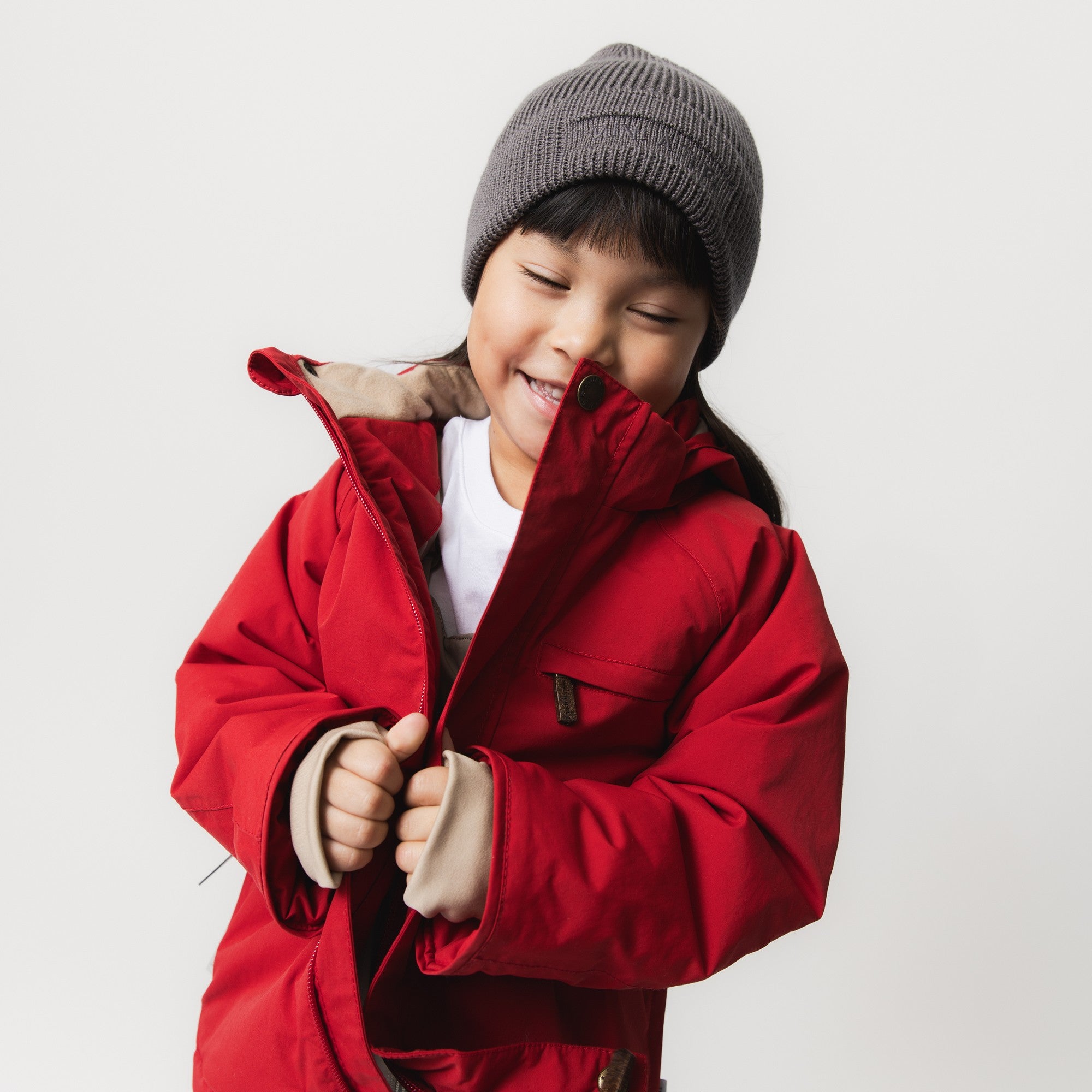Young girl wearing the MATBRIDDINE winter jacket in red with a grey knitted beanie, smiling while adjusting the front zip, showing the beige inner lining and snow cuffs with thumb openings at the wrists.