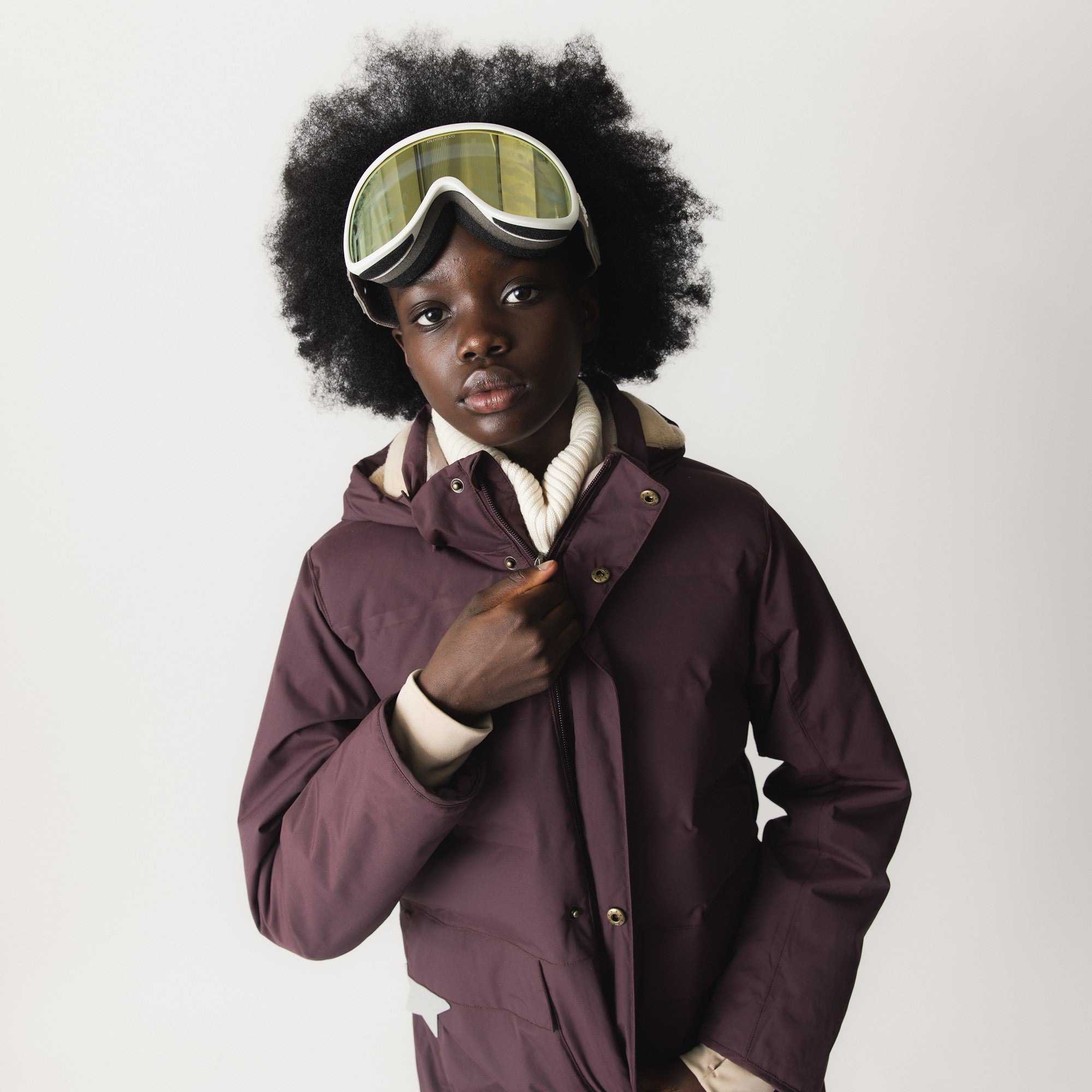 MATVENCASIA winter jacket worn with ski goggles, showing the snow cuffs with cream-coloured thumb openings at the wrists, detachable hood with snap-button fastening, and the waterproof shell in a deep burgundy tone.