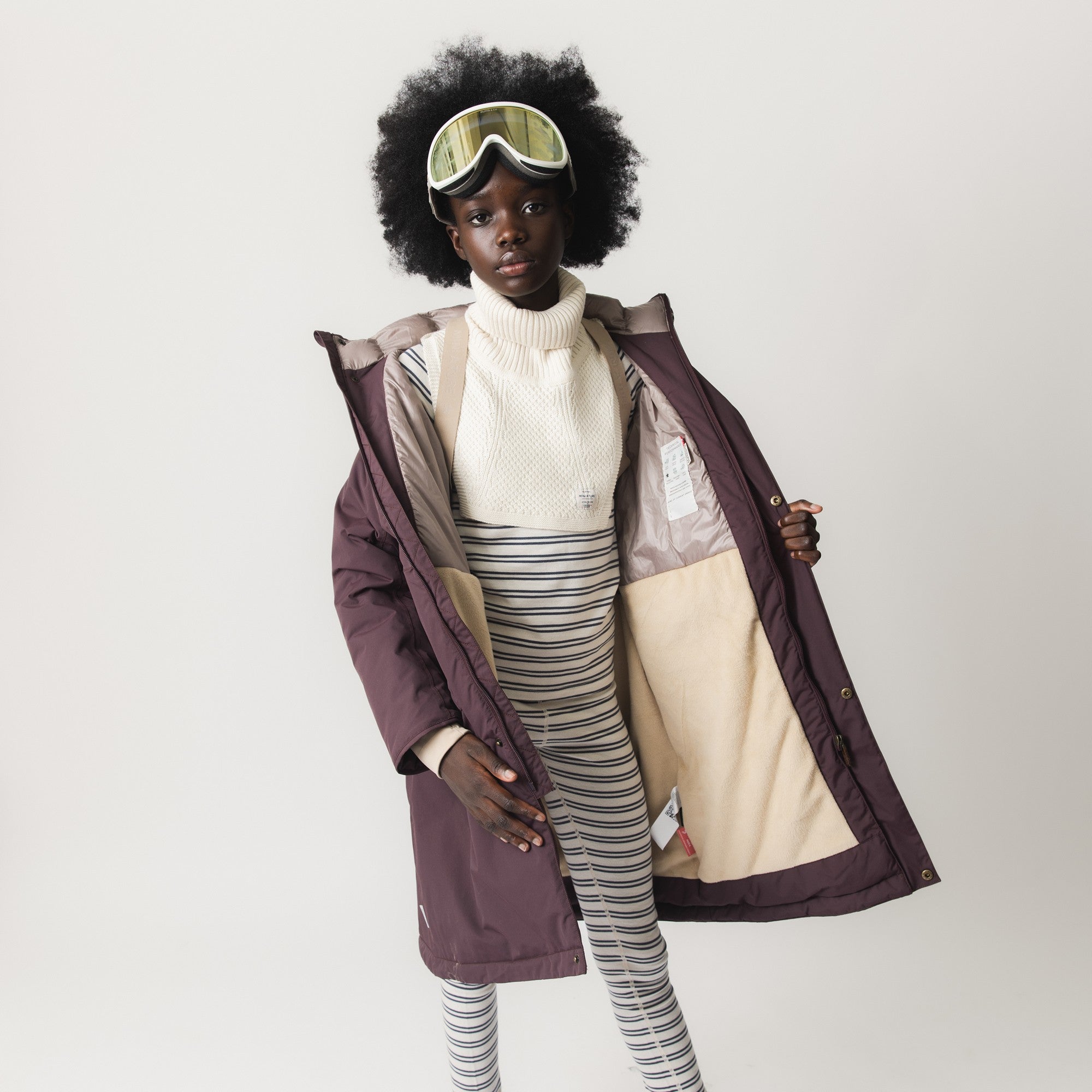 MATVENCASIA winter jacket held open to display the internal suspenders in beige, the Sorona Aura padded upper section, the soft fleece lining in the lower half, and the snap-button hood attachment, worn over a cream turtleneck and striped trousers.