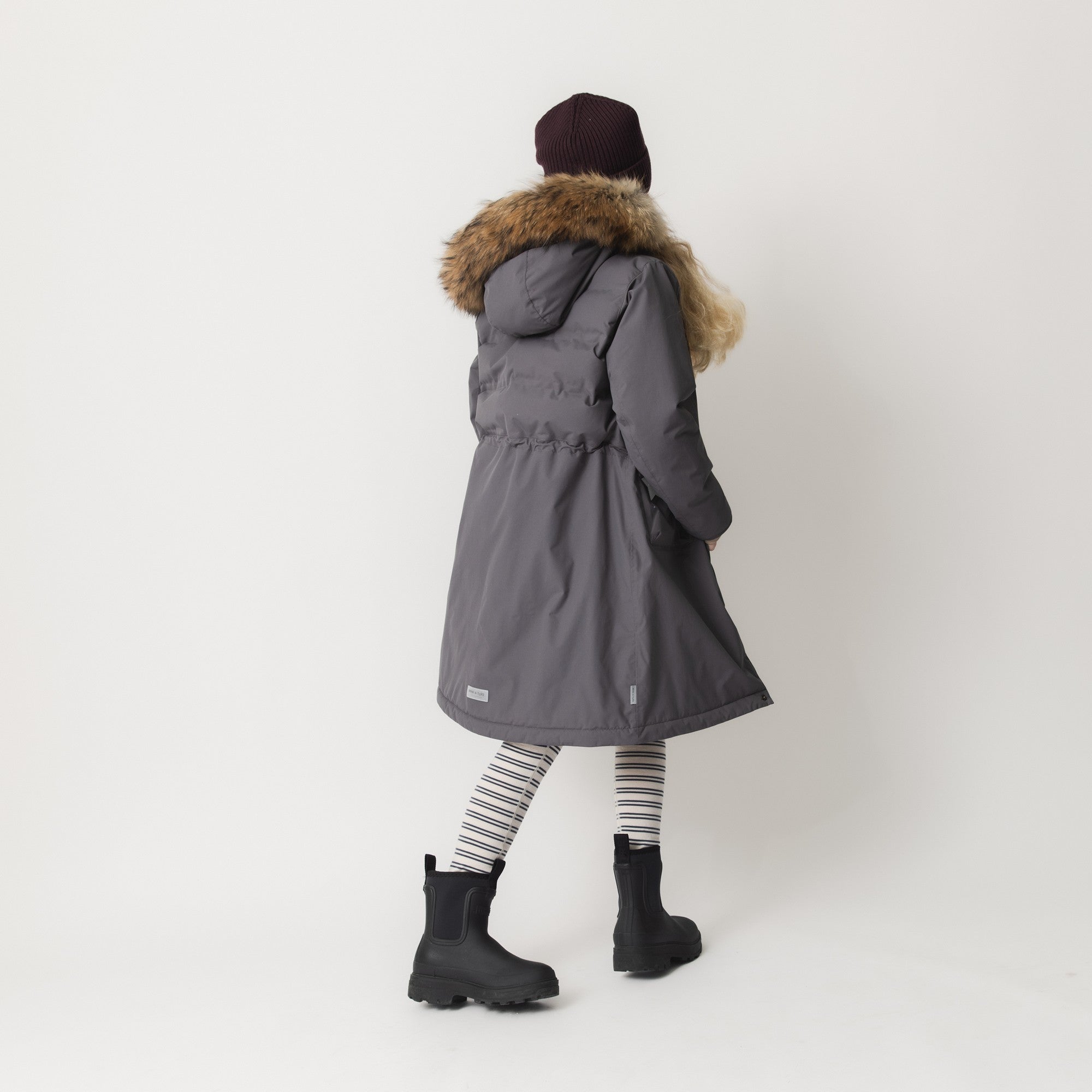 MATVENCASIA winter jacket worn by a girl seen from behind, highlighting the extended length that reaches mid-thigh, the padded upper body transitioning to a fleece-lined lower section, and the detachable hood with genuine fur trim framing the shoulders.