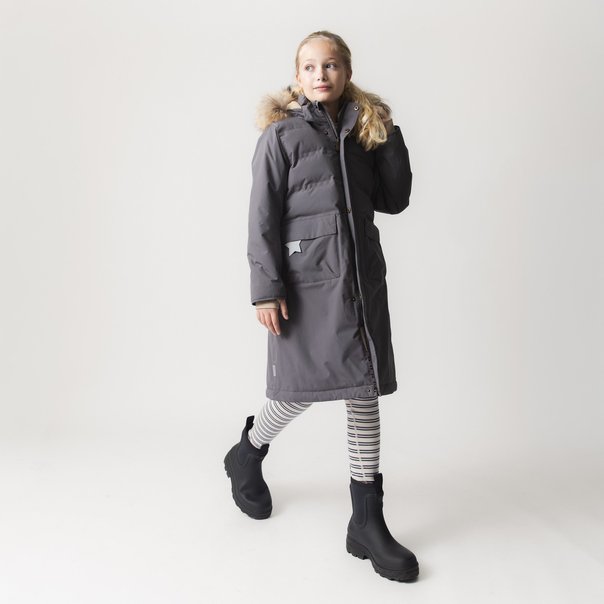 MATVENCASIA fleece lined winter jacket in dark grey, worn by a girl with striped leggings and black boots, showing the longer cut that extends below the waist and the detachable hood with real fur trim.