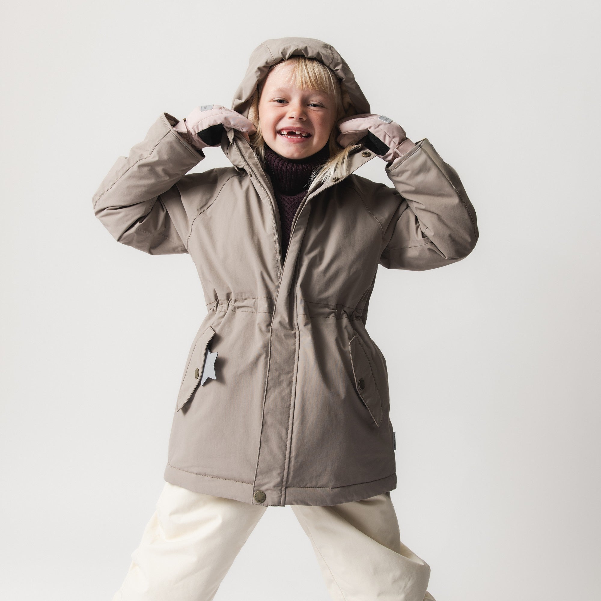 MATVIKANIA fleece lined winter jacket in taupe with detachable hood, worn by a smiling child demonstrating the defined waist and extended length silhouette.