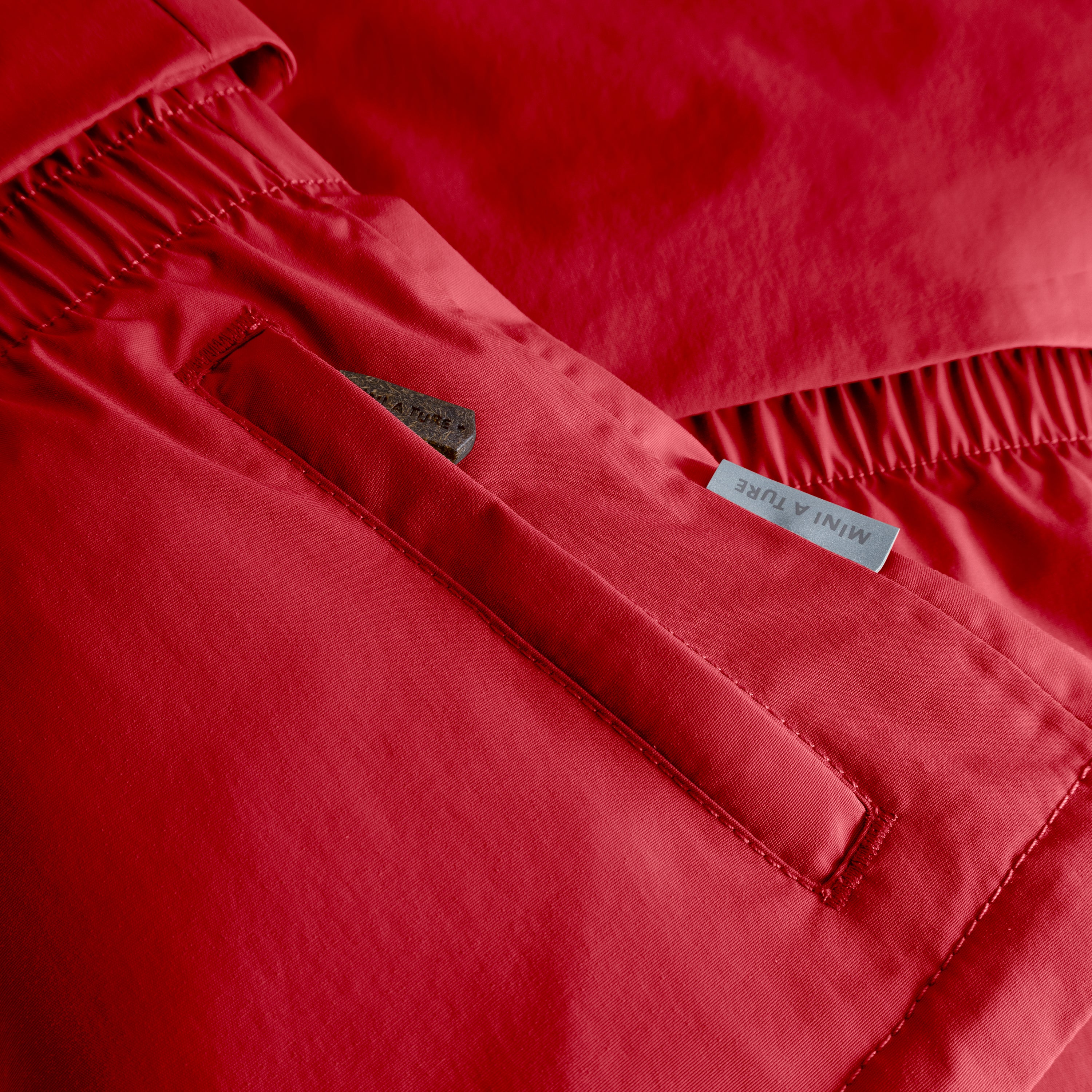 MATALGEANA winter jacket detail in red showing the internal snow cuff with thumb opening at the sleeve, beige fleece lining visible at the cuff edge, elasticated waist panel with gathered stitching, and MINI A TURE woven label on the waterproof recycled polyamide shell.