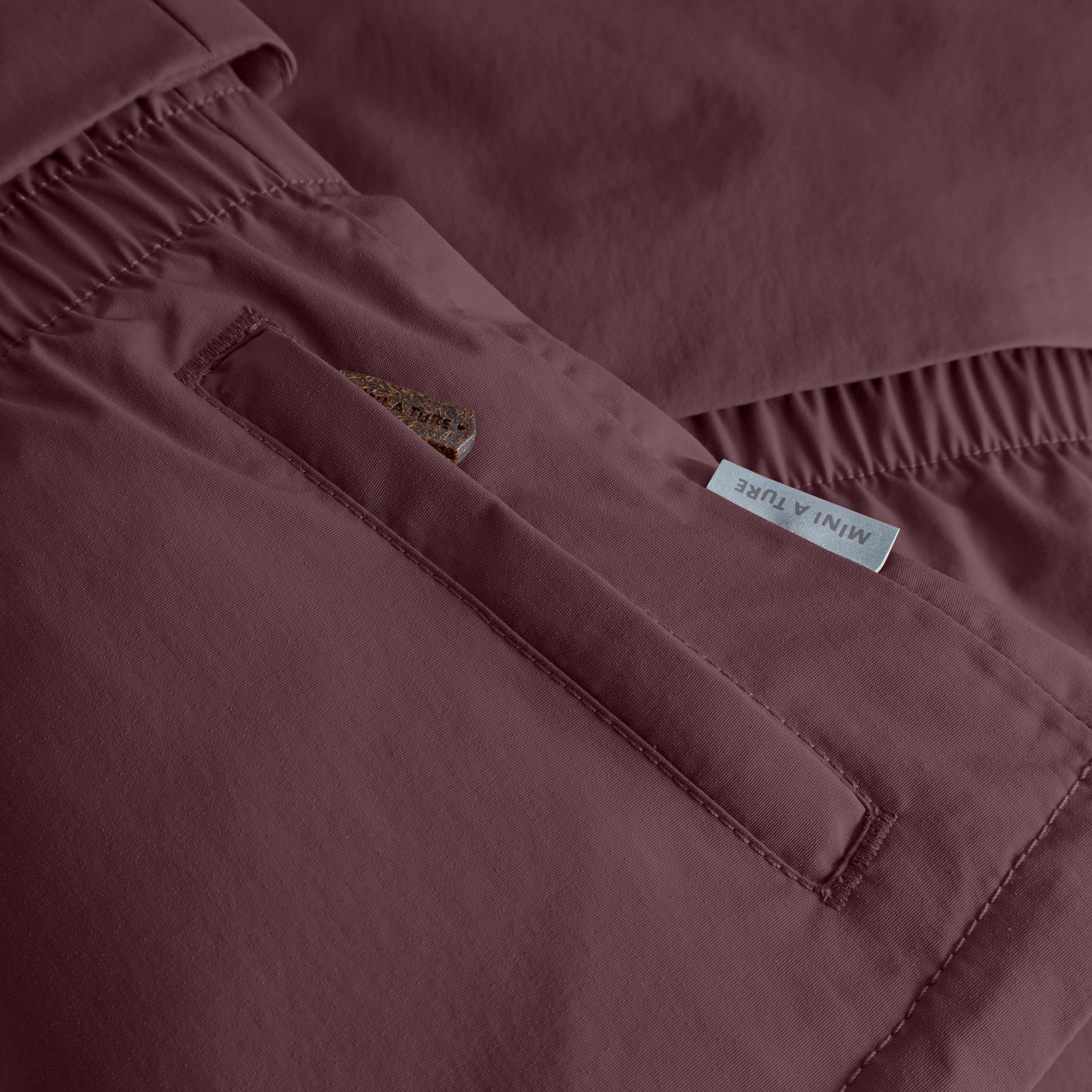 MATALGEANA winter jacket detail showing the right pocket with a 70 lux reflective star emblem and MINI A TURE label, alongside the elasticated waist panel and recycled polyamide shell in burgundy.