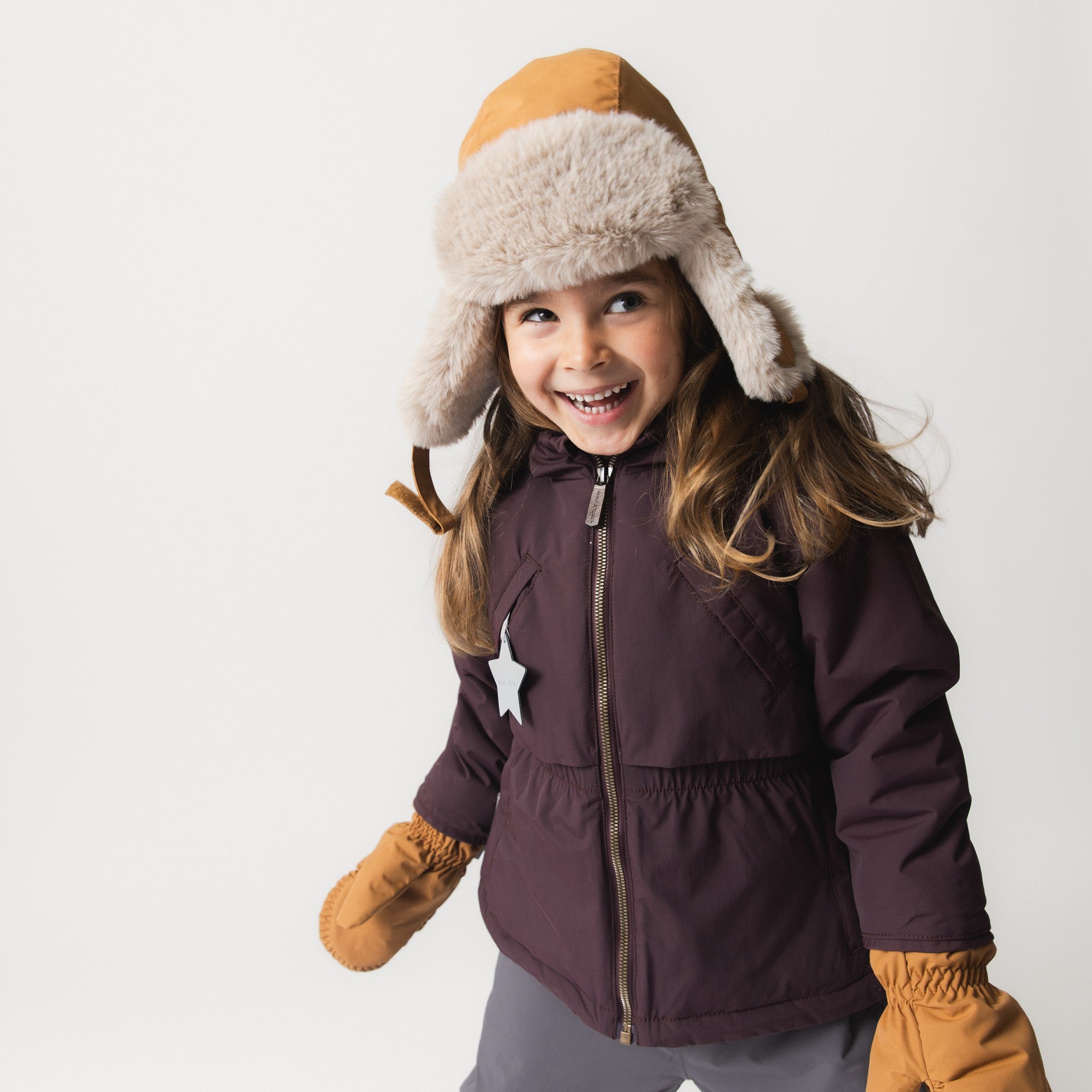 MATALGEANA fleece lined winter jacket in deep plum with a trench-inspired silhouette, featuring a cape detail at the shoulders, fixed elastic waist, and reflective star detail on the pocket, worn by a smiling girl with a fur-trimmed hat and mustard mittens.