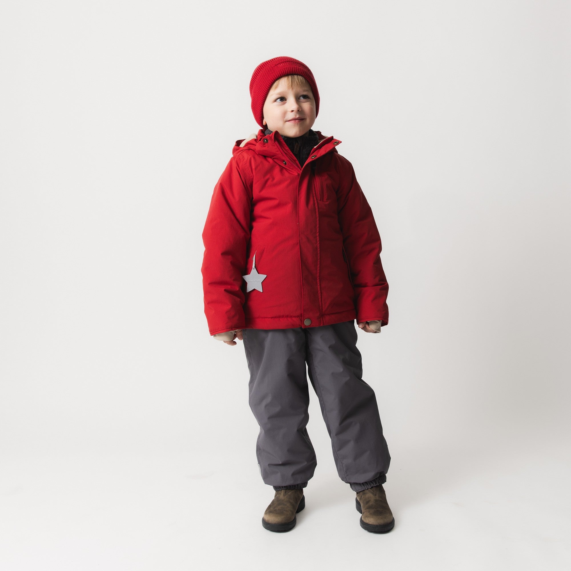 Child wearing MATADYANKO fleece lined winter jacket in red with detachable hood, paired with grey trousers and a red knitted beanie, showing the jacket's regular fit and reflective star detail.