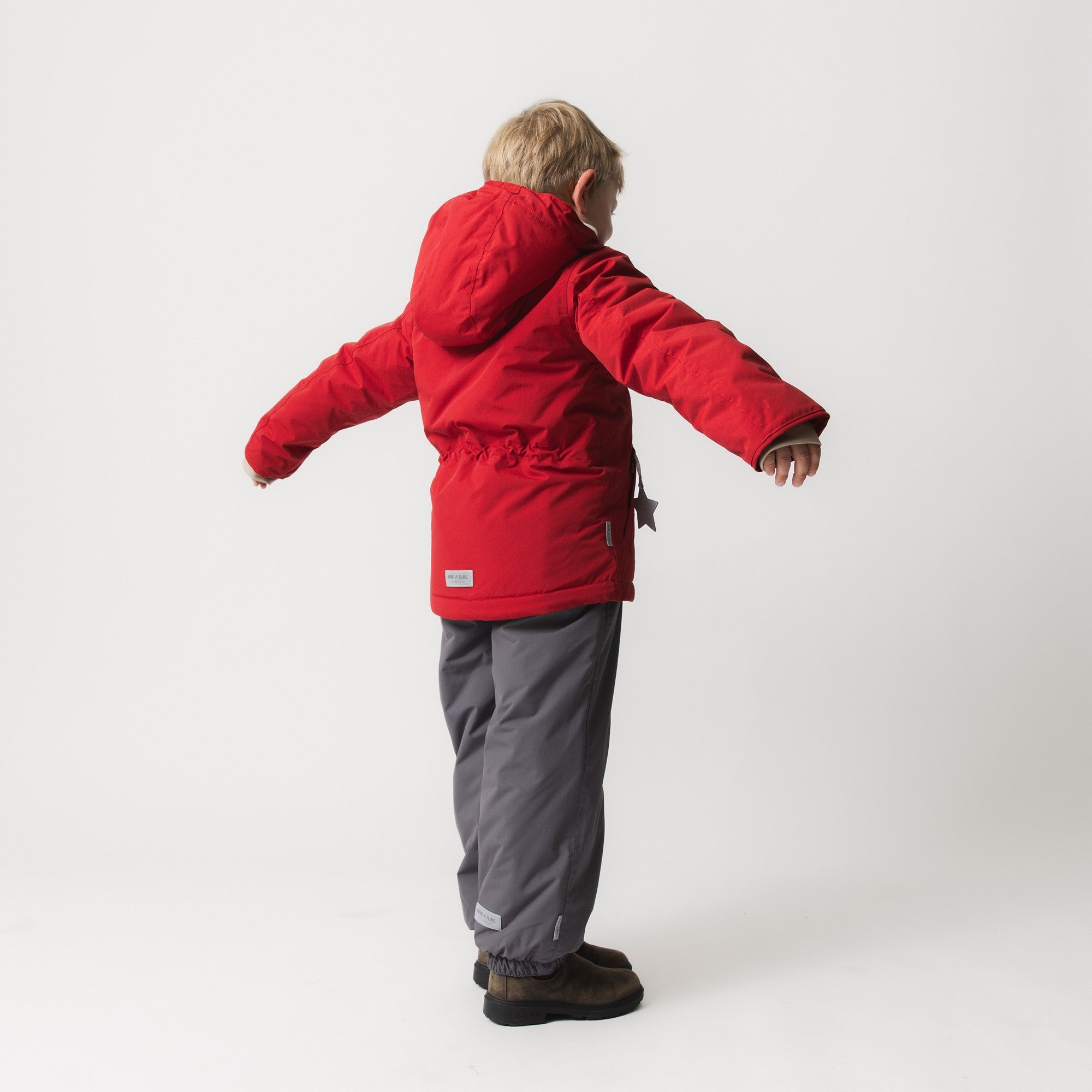 Child seen from behind wearing MATADYANKO fleece lined winter jacket in red with arms outstretched, demonstrating the improved sleeve shaping and freedom of movement, paired with grey trousers.