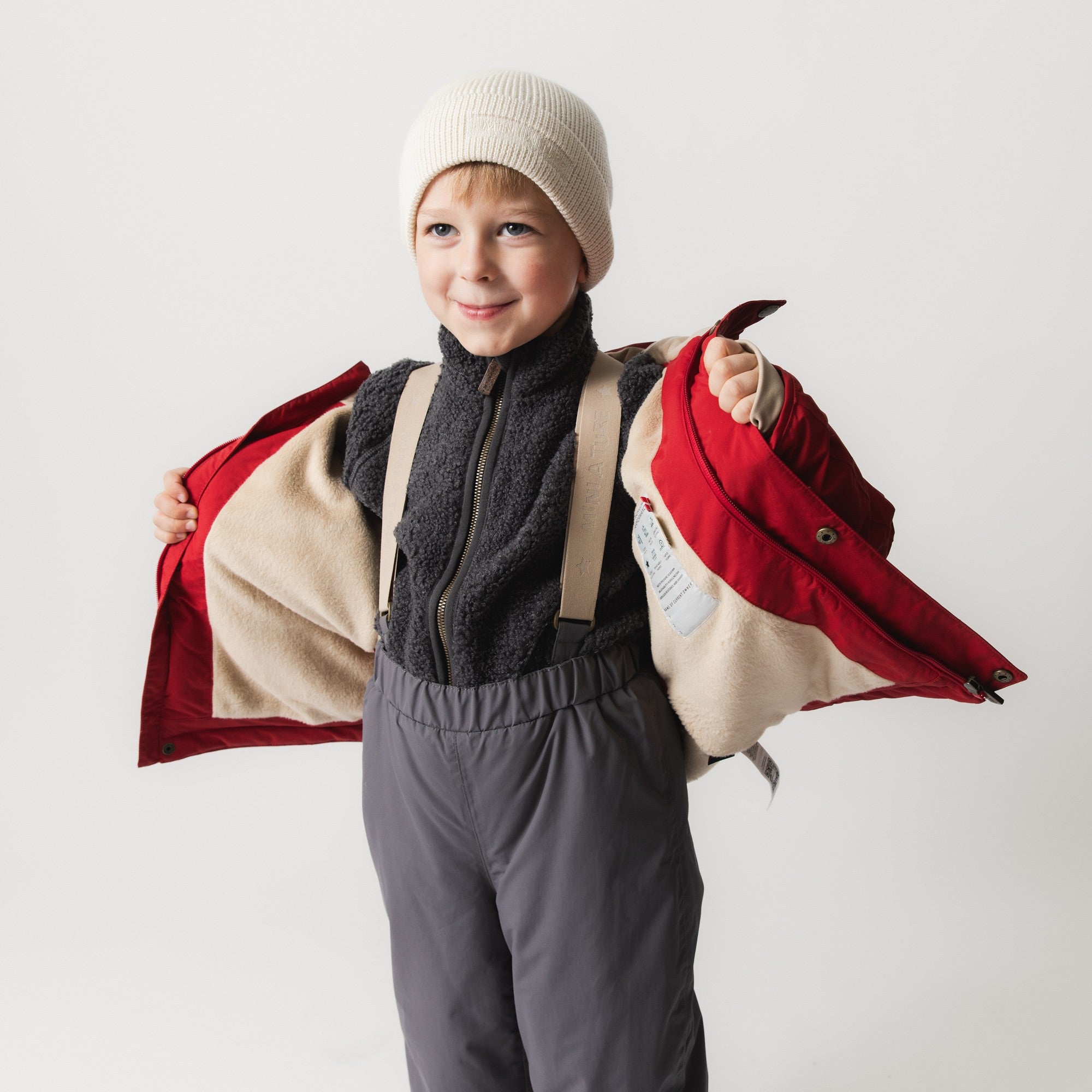 Child holding MATADYANKO fleece lined winter jacket open to reveal the soft beige fleece lining and internal construction, layered over a black fleece mid-layer and grey trousers, wearing a cream knitted beanie.