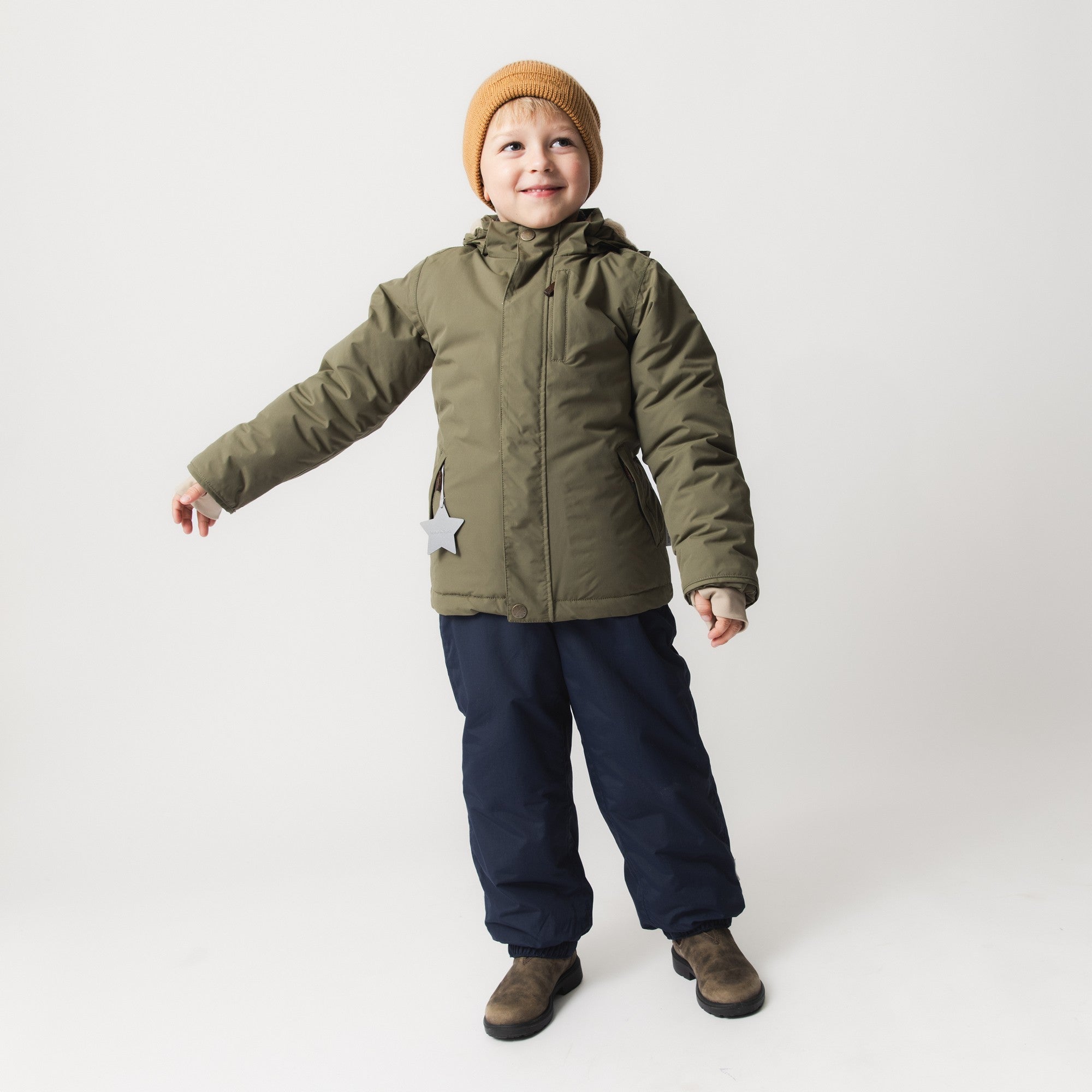 Child wearing MATADYANKO fleece lined winter jacket in olive green with detachable hood, paired with navy insulated trousers, brown boots and a caramel knitted beanie, demonstrating the jacket's regular fit and freedom of movement during active play.