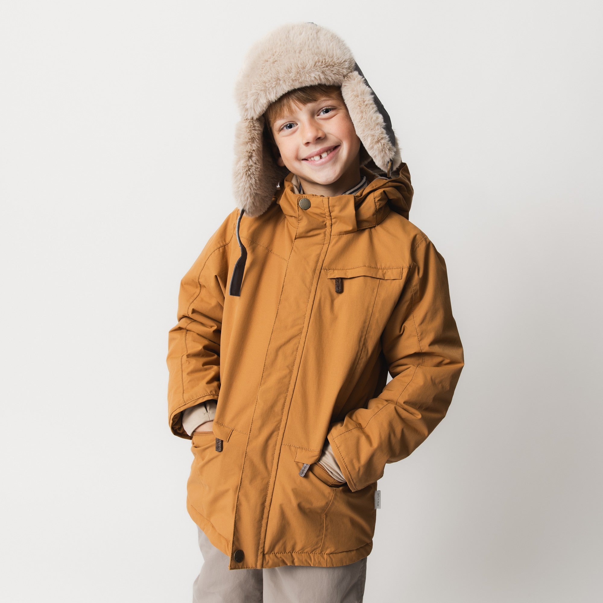 MATVESTYN winter jacket in caramel brown worn by a smiling child, featuring a detachable faux-fur trimmed hood, front zip closure with snap-button placket, and front pockets with hidden zips.