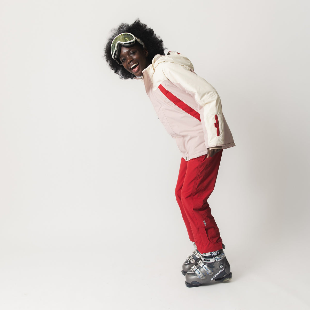 MATVALONSO Skijacke in Creme und Rosa mit roten Streifen, getragen von einem Kind mit Skibrille und Skischuhen, zeigt die verbesserten Ärmelschnitte und Schneemanschetten mit Daumenloch.