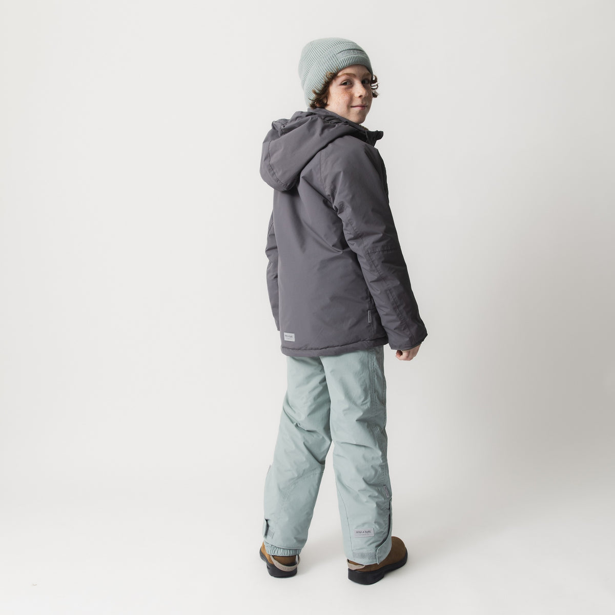 MATVALONSO Skijacke in Grau mit abnehmbarer Kapuze, getragen von einem Kind mit grauer Strickmütze und schwarzem Schal, kombiniert mit mintgrüner Skihose und braunen Schuhen. Die Rückansicht zeigt die normale Passform und die vollständig versiegelten Nähte.