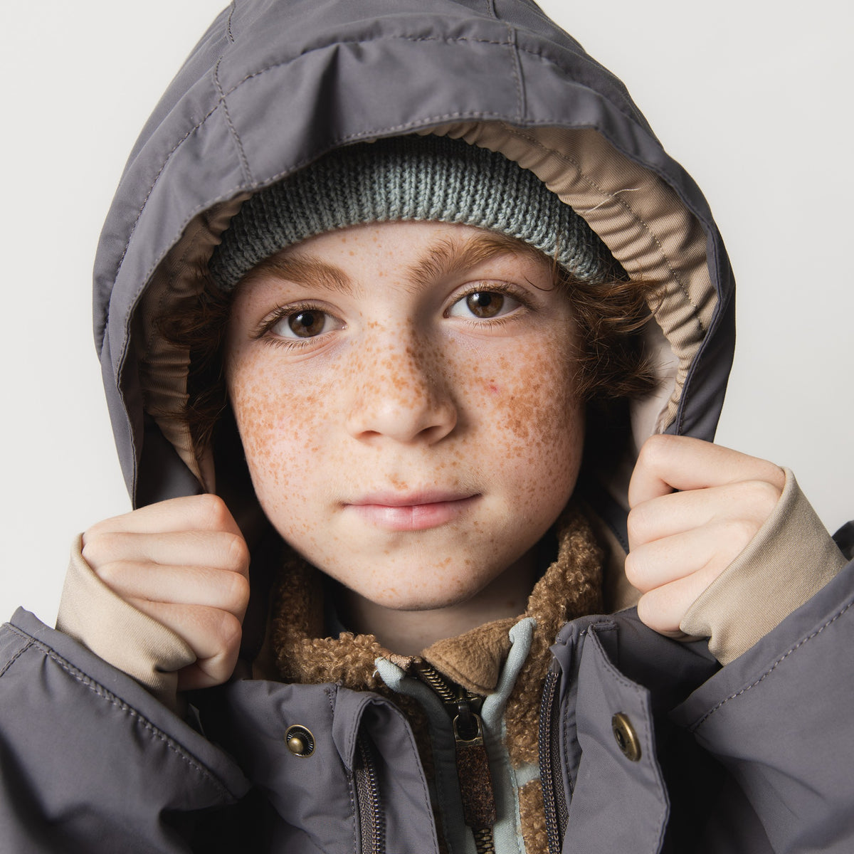 MATVALONSO Skijacke in Grau mit abnehmbarer Kapuze, getragen von einem Kind mit grüner Strickmütze und braunem Schal, zeigt die Nahaufnahme der Kapuze mit cremefarbenem Innenfutter und Druckknopfbefestigung.