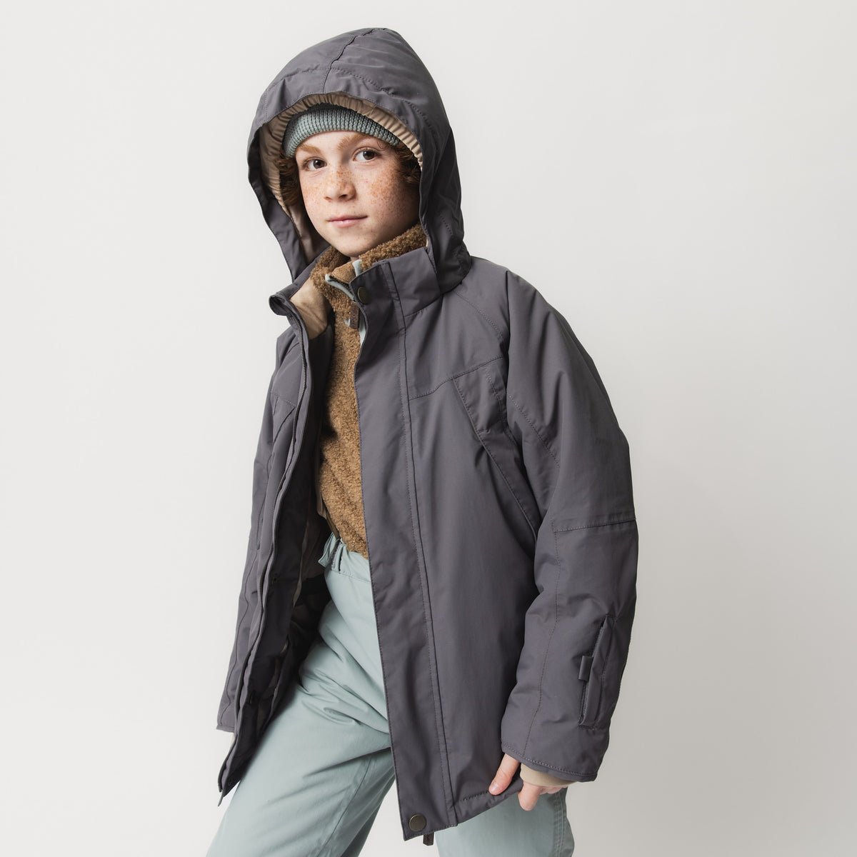 MATVALONSO Skijacke in Grau mit abnehmbarer Kapuze, getragen von einem Kind mit gestrickter Mütze und braunem Schal, kombiniert mit hellblauer Skihose, zeigt die normale Passform und die verbesserten Ärmelschnitte.