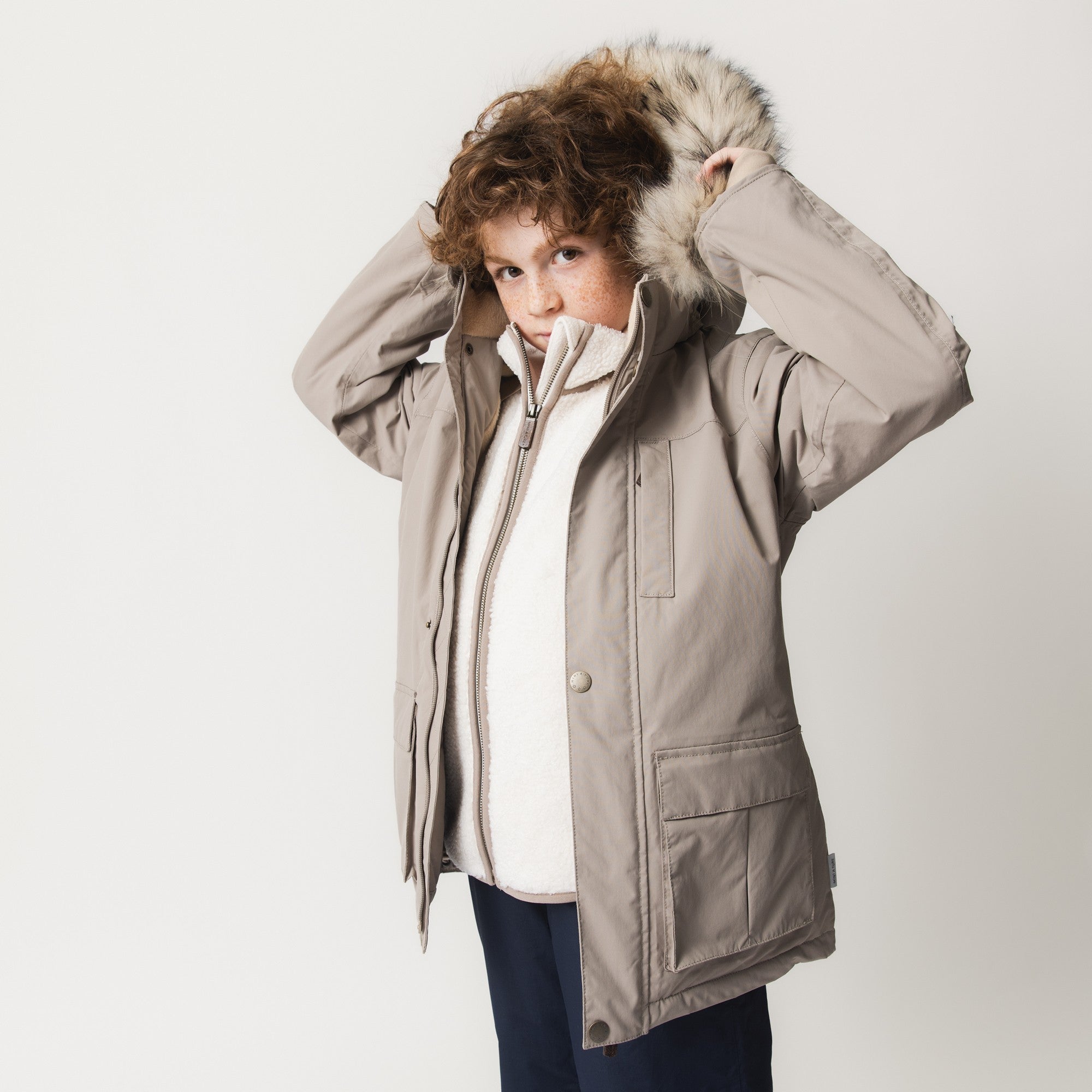 MATKASTORIO fleece lined winter jacket in beige, three-quarter front view worn by a child adjusting the detachable hood with genuine fur trim, highlighting the snap button fastening, front zip closure and front pockets with magnetic closures.