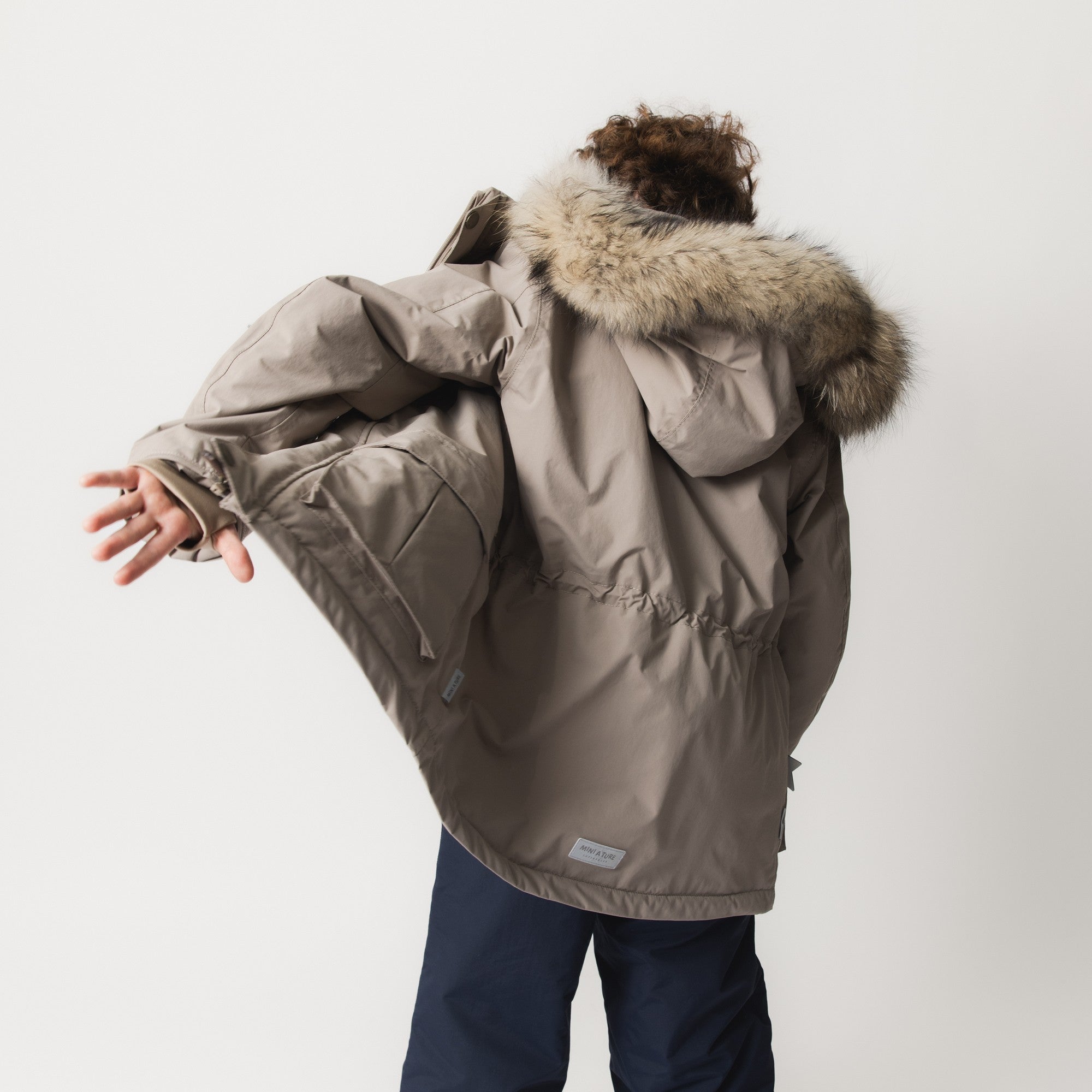 MATKASTORIO fleece lined winter jacket in beige, side view worn by a child with arms extended, showing the improved sleeve fit, snow cuffs at the wrists, detachable hood with genuine fur trim and the waterproof recycled polyamide shell construction.