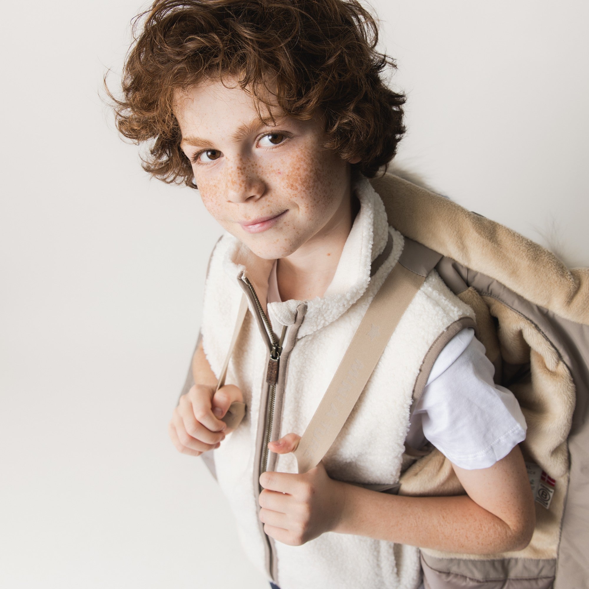 MATKASTORIO fleece lined winter jacket in beige draped over a child's shoulder, showing the soft fleece lining at the collar, front zip closure and the jacket's utility as a layering piece that can be worn loosely when indoor spaces become too warm.
