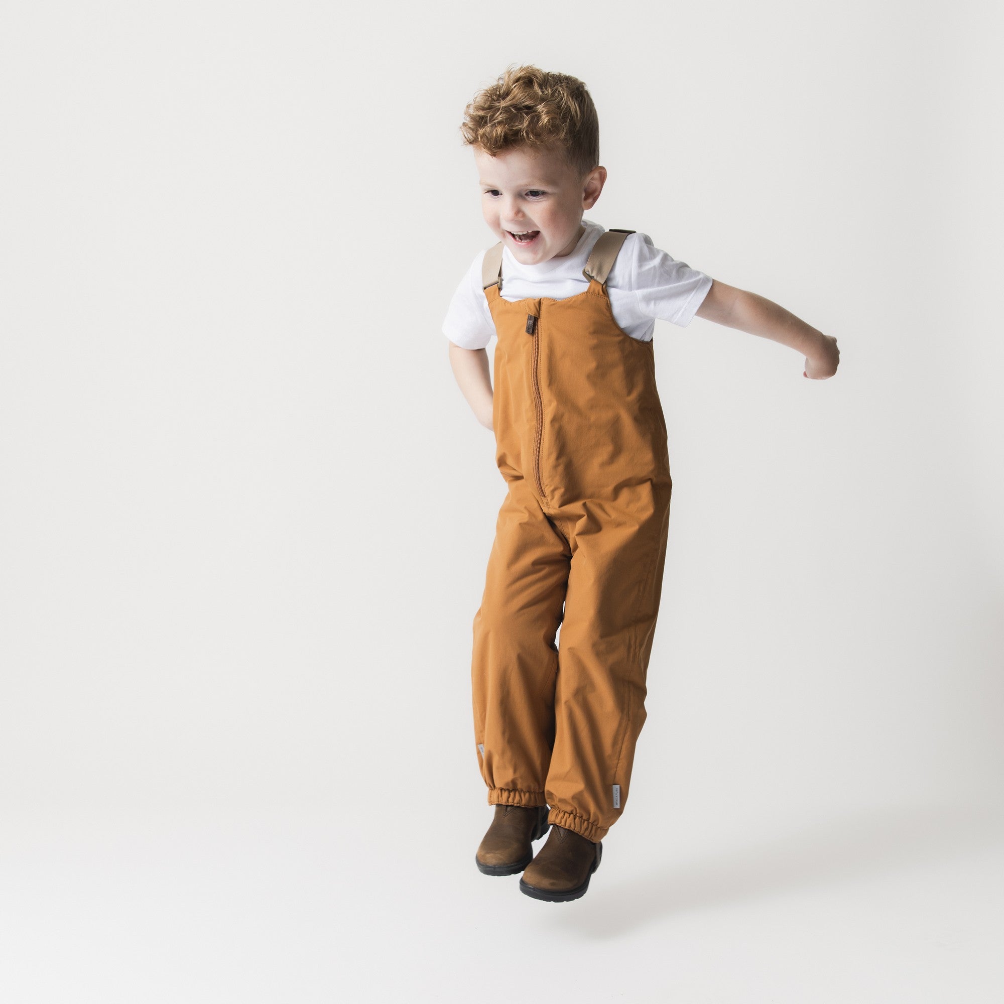 MATWALENTEX snow pants in warm caramel worn by a smiling toddler over a white t-shirt, showing the full-length front zip, adjustable suspenders and elasticated ankle cuffs with foot straps.