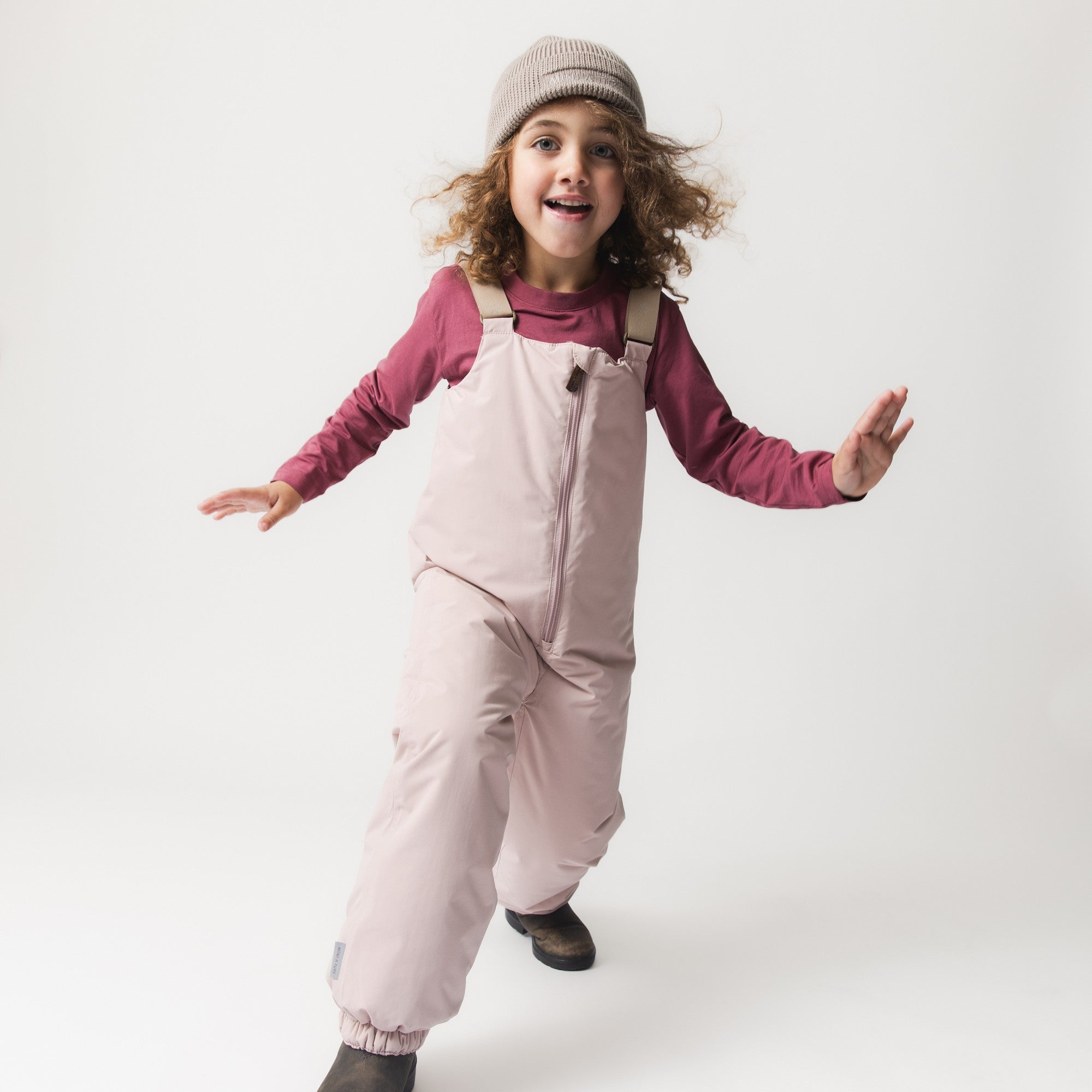 MATWALENTEX snow pants in soft pink worn by a child mid-jump, illustrating the dropped crotch design and unrestricted movement, paired with a berry-coloured long-sleeve top, beige suspenders and a knitted beanie.