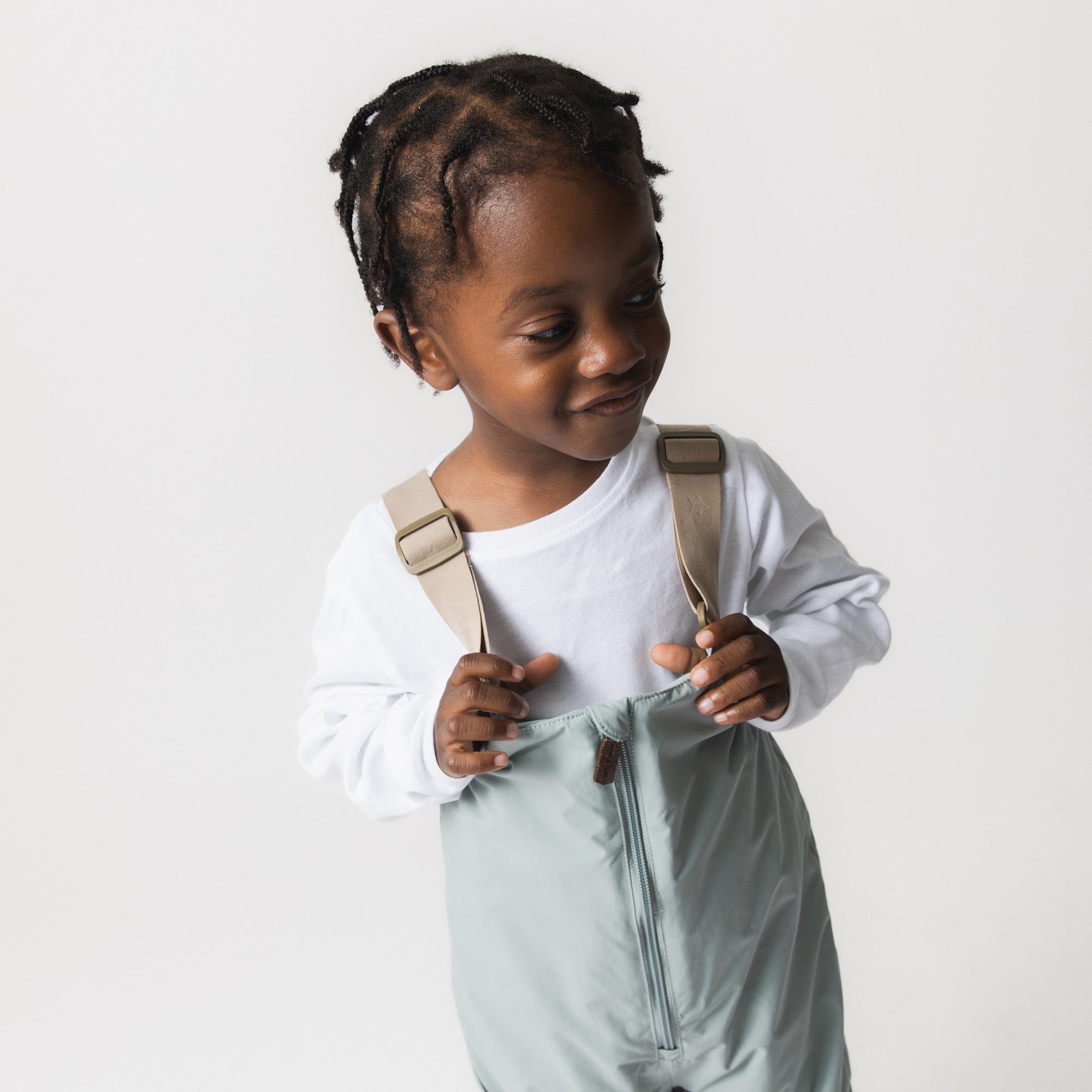 MATWALENTEX snow pants in pale mint green worn by a toddler adjusting the beige suspenders, paired with a white long-sleeve top, demonstrating the front zip opening and relaxed fit through the waist and legs.