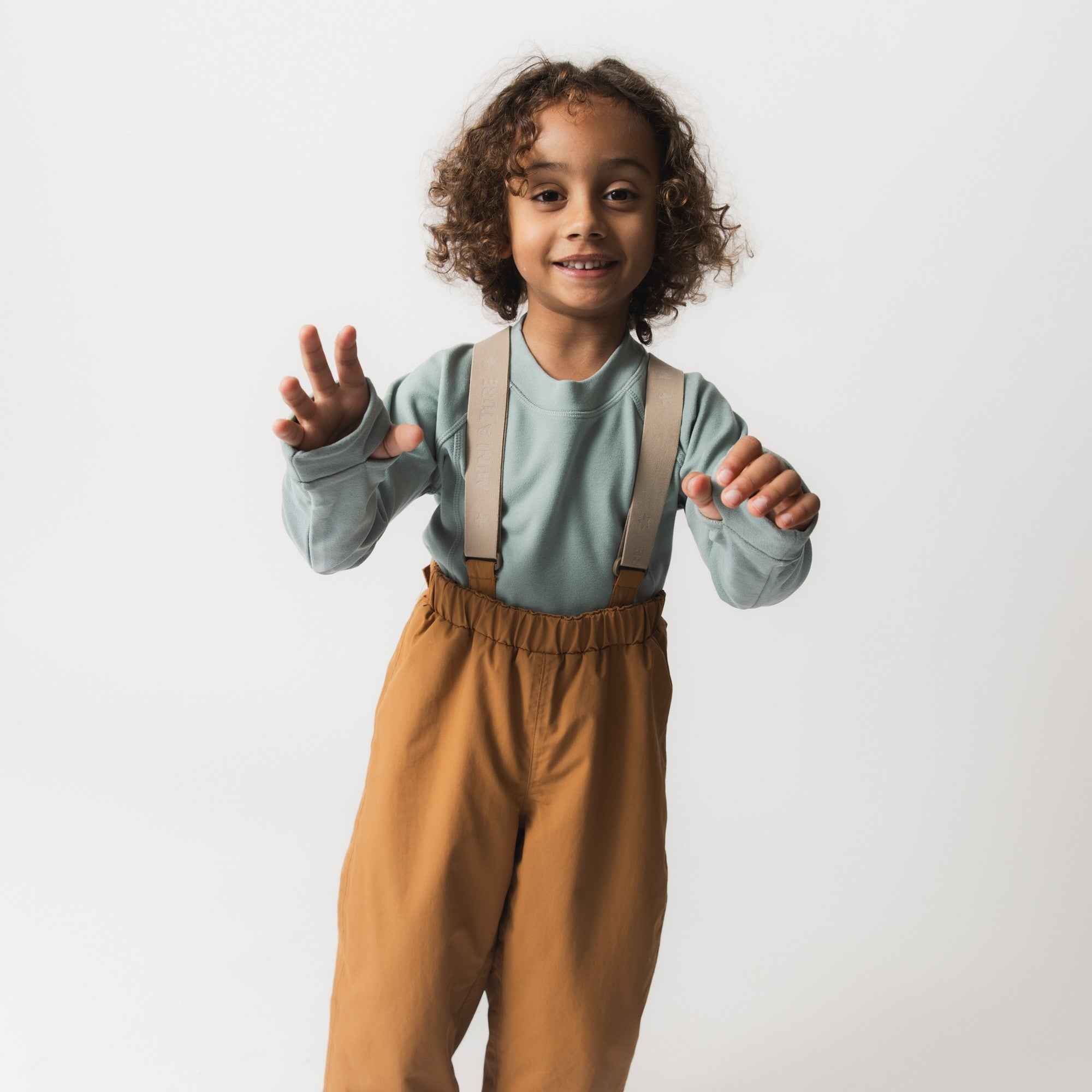 Child wearing MATWILAS snow pants in golden caramel with beige adjustable braces, paired with a sage green long-sleeve top, standing in a playful pose with both hands raised.