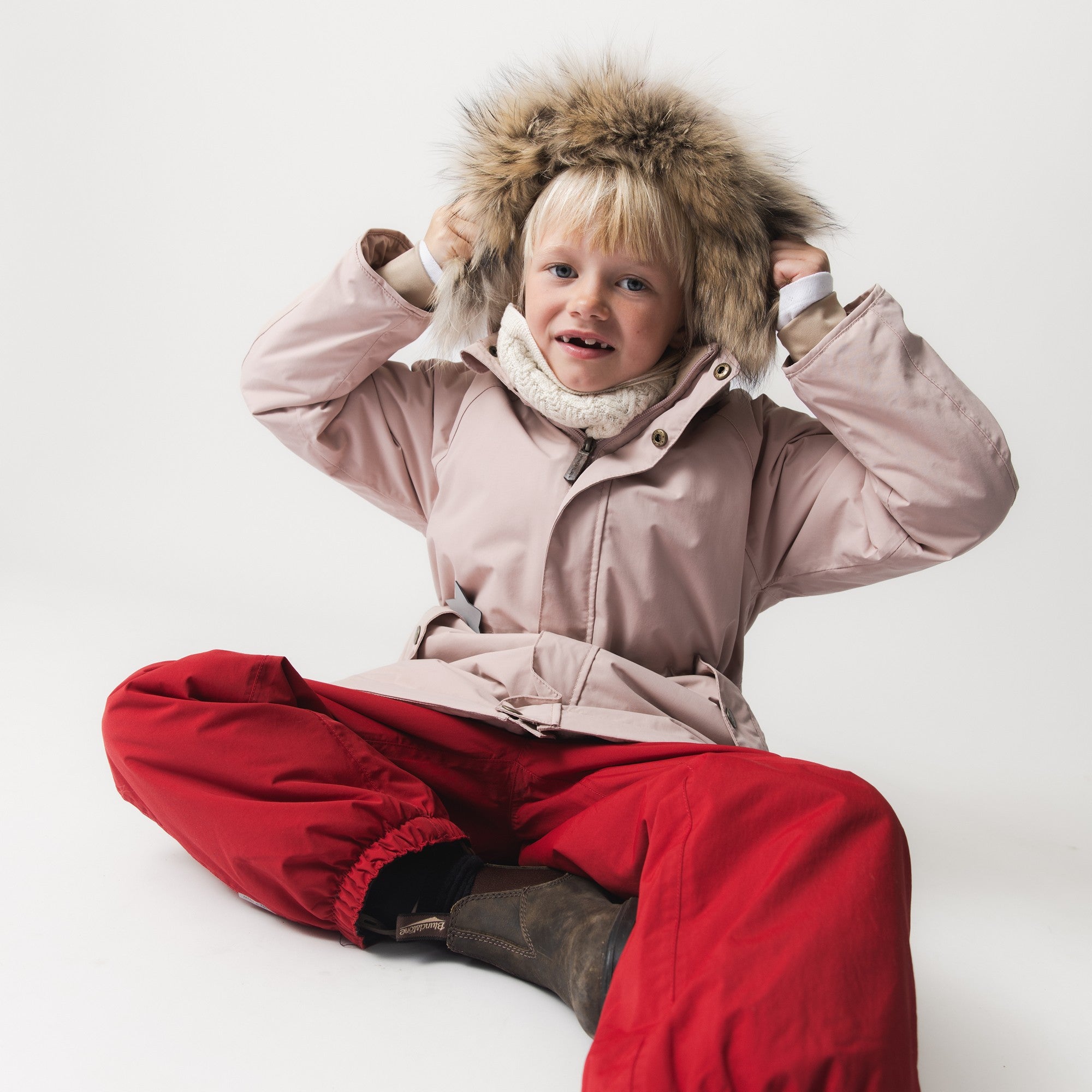 Child seated wearing MATWILAS snow pants in bright red with elasticated ankle cuffs, paired with a pale pink winter jacket featuring a fur-trimmed hood and cream knitted neck warmer.