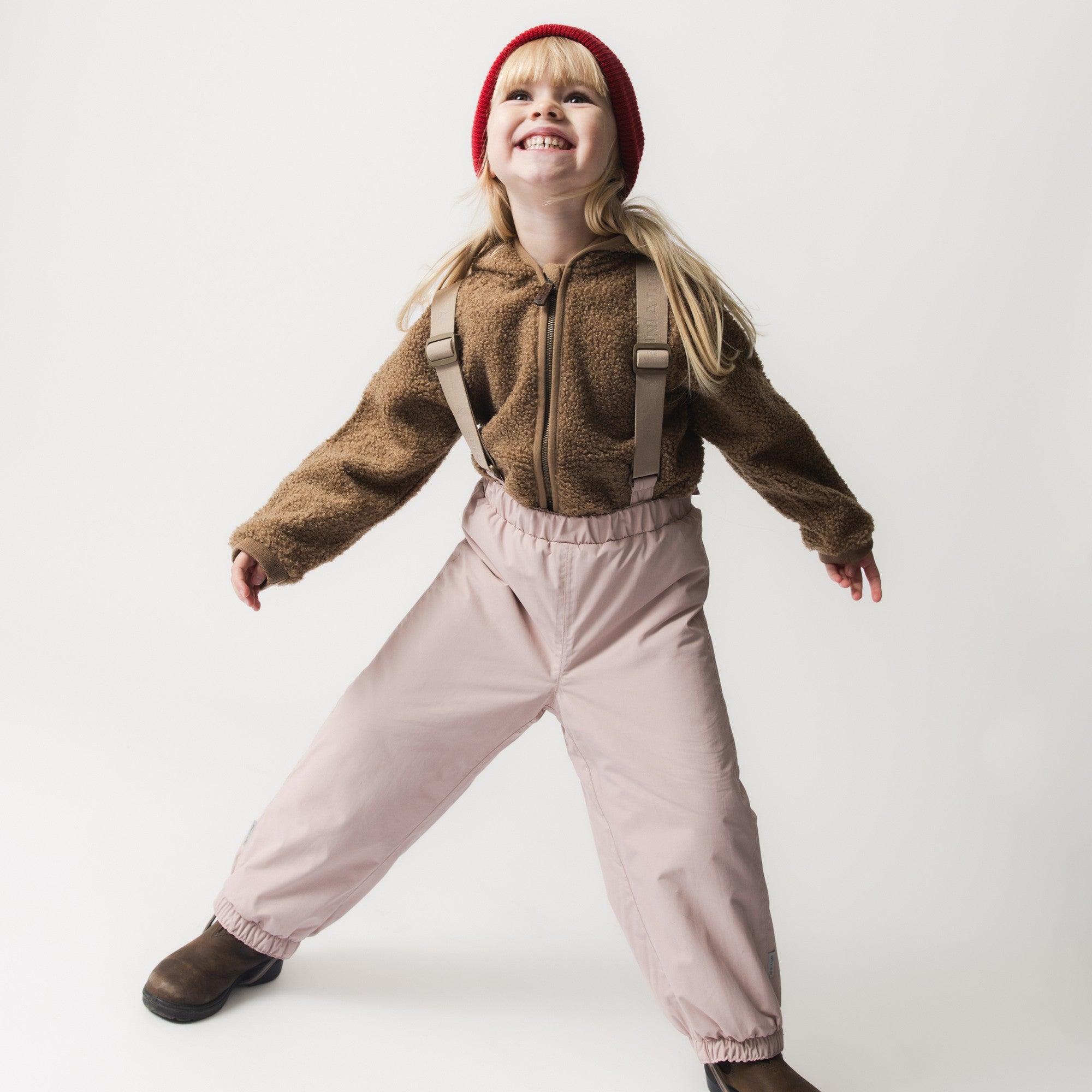 Child wearing MATWILAS snow pants in soft pink with beige adjustable braces, paired with a brown teddy fleece jacket and red knitted beanie, standing in an active pose.