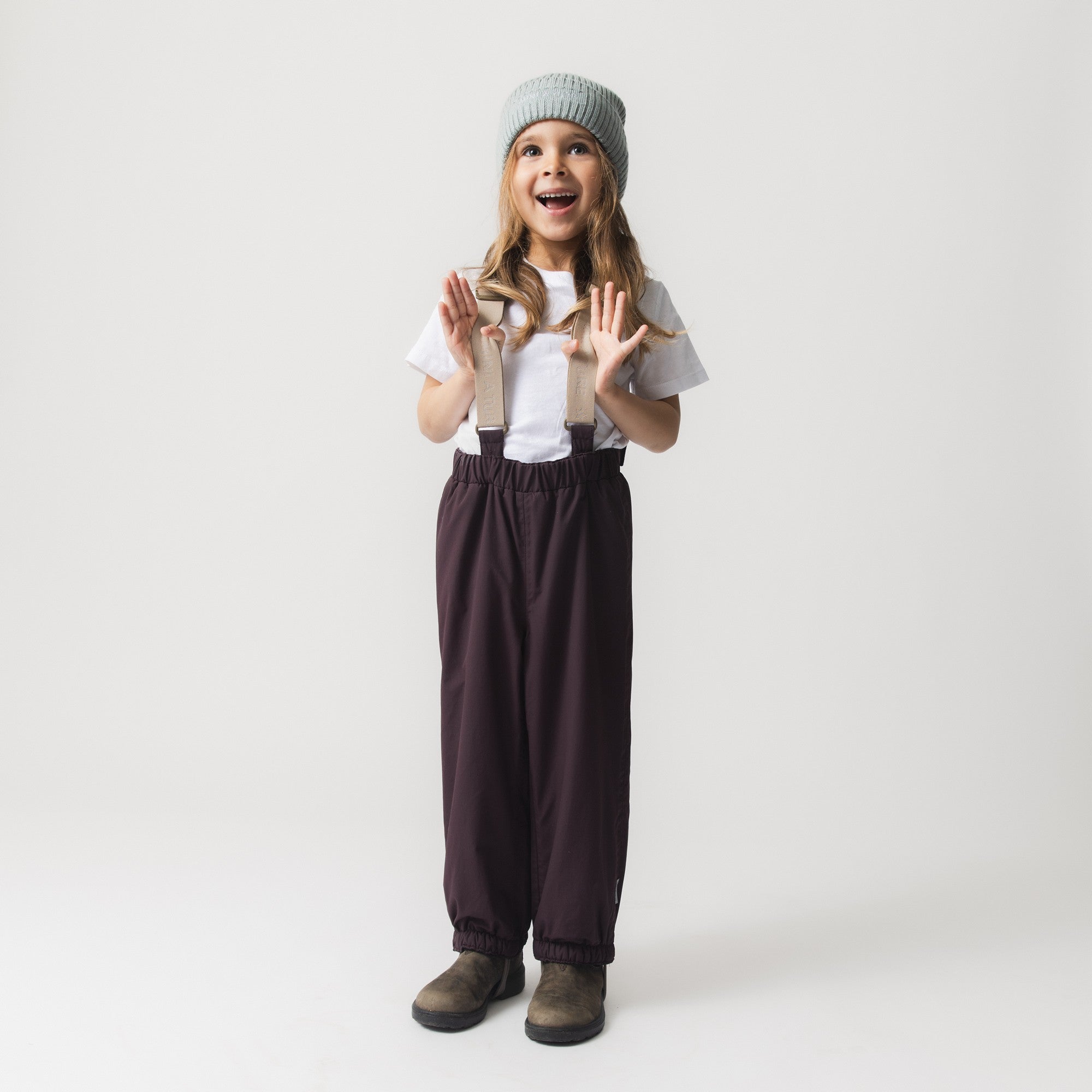 Child wearing MATWILAS snow pants in deep plum with beige adjustable braces, paired with a white t-shirt and grey knitted beanie, standing with hands raised in a joyful expression.