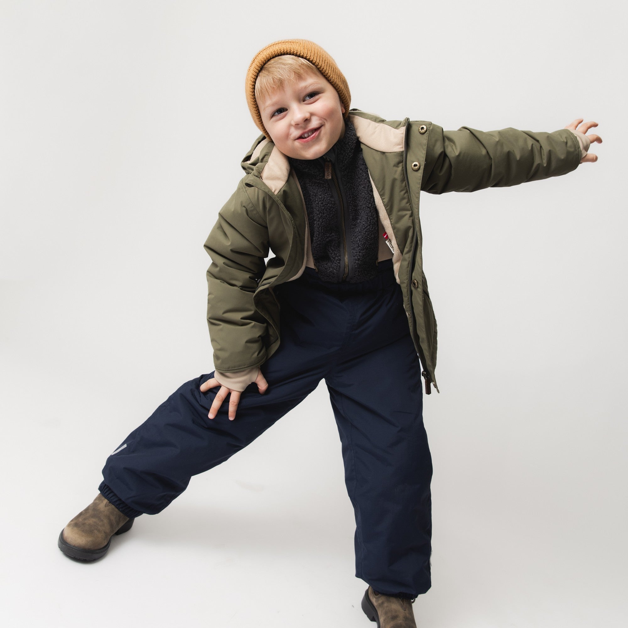 Child wearing MATWILAS snow pants in navy blue with elasticated ankle cuffs, styled with an olive green winter jacket, black fleece layer and camel knitted beanie, posing playfully in a studio setting.
