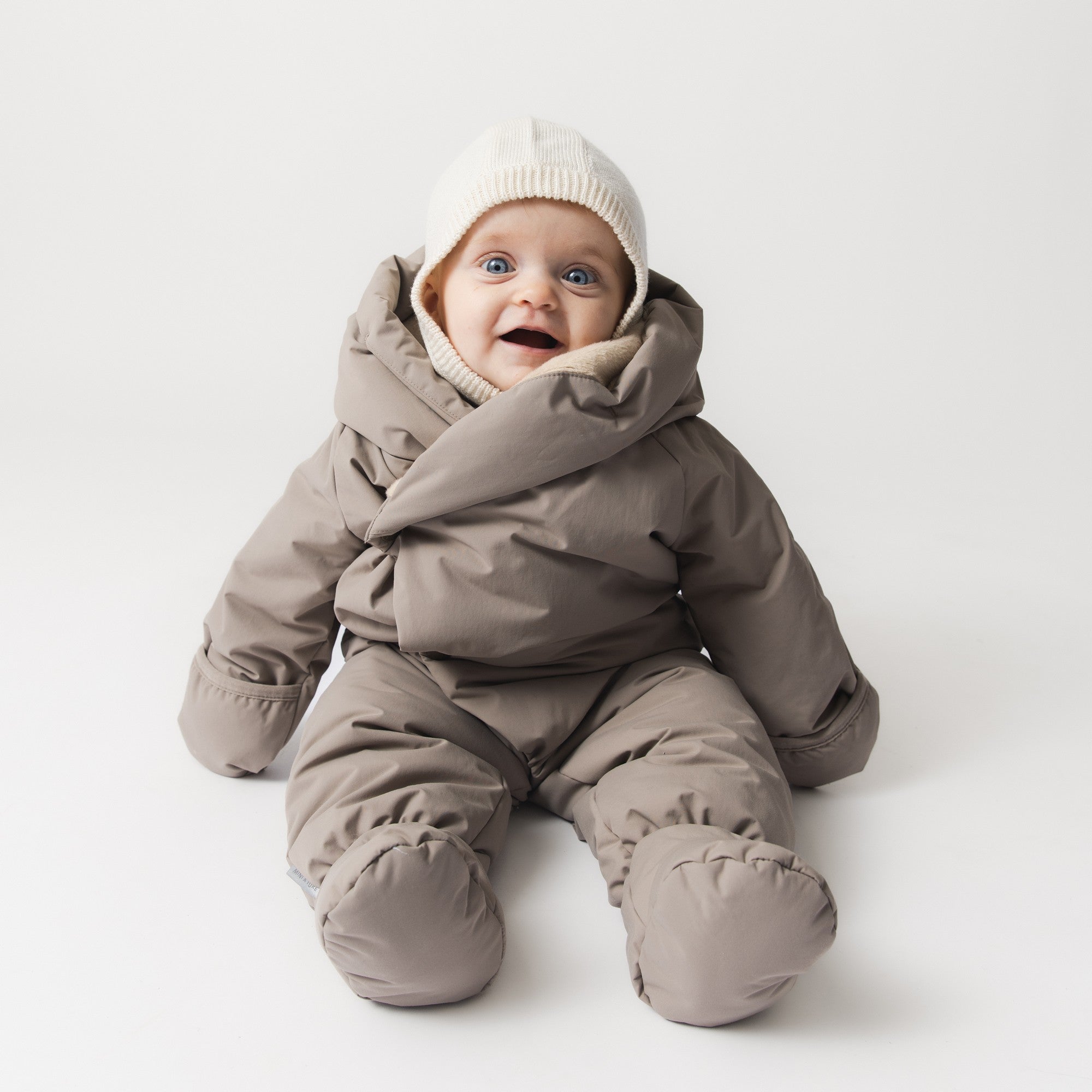 MATFIANNA fleece lined winter pramsuit worn by a smiling baby in a seated position, showing the shawl-collar hood framing the face, THERMOLITE padded body and sleeves, and integrated socks in taupe grey waterproof recycled polyamide.