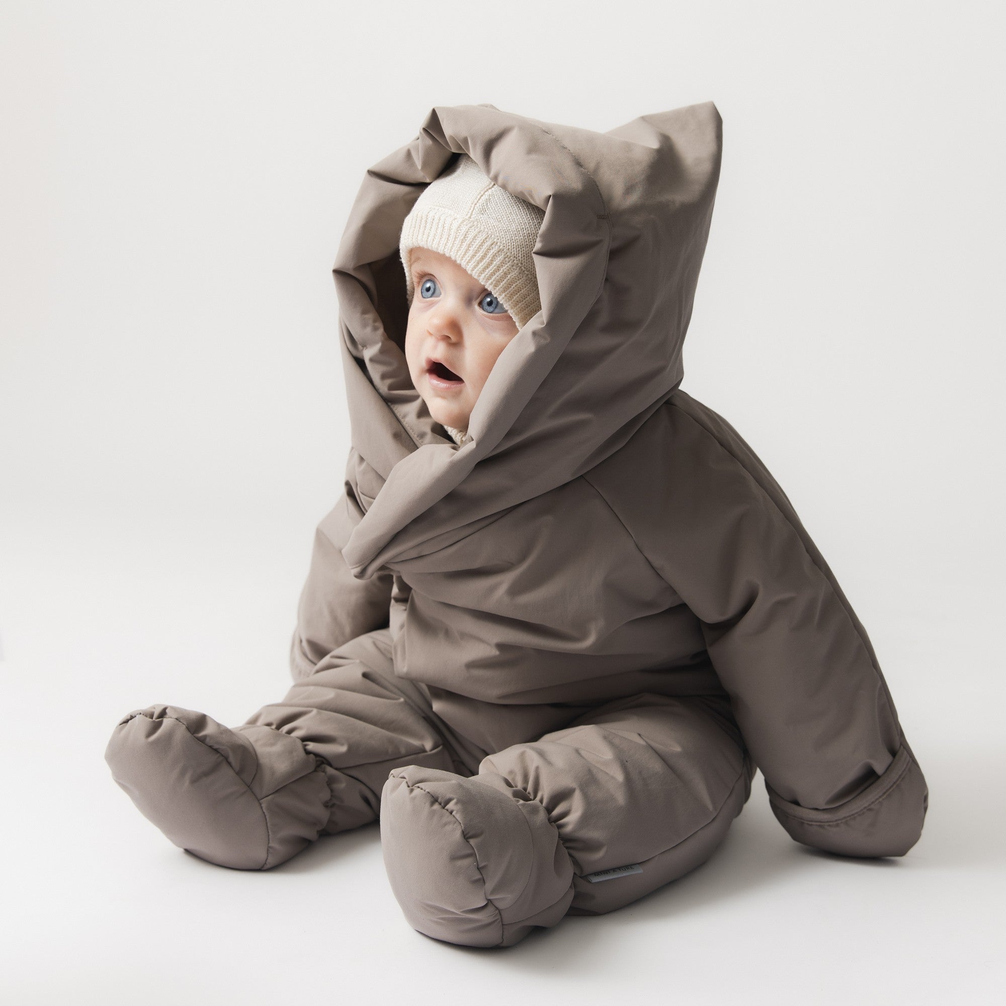 MATFIANNA fleece lined winter pramsuit in taupe grey, shown on a seated baby wearing a cream knitted hat, with shawl-collar hood, built-in socks and fold-over mittens for full-body winter coverage.