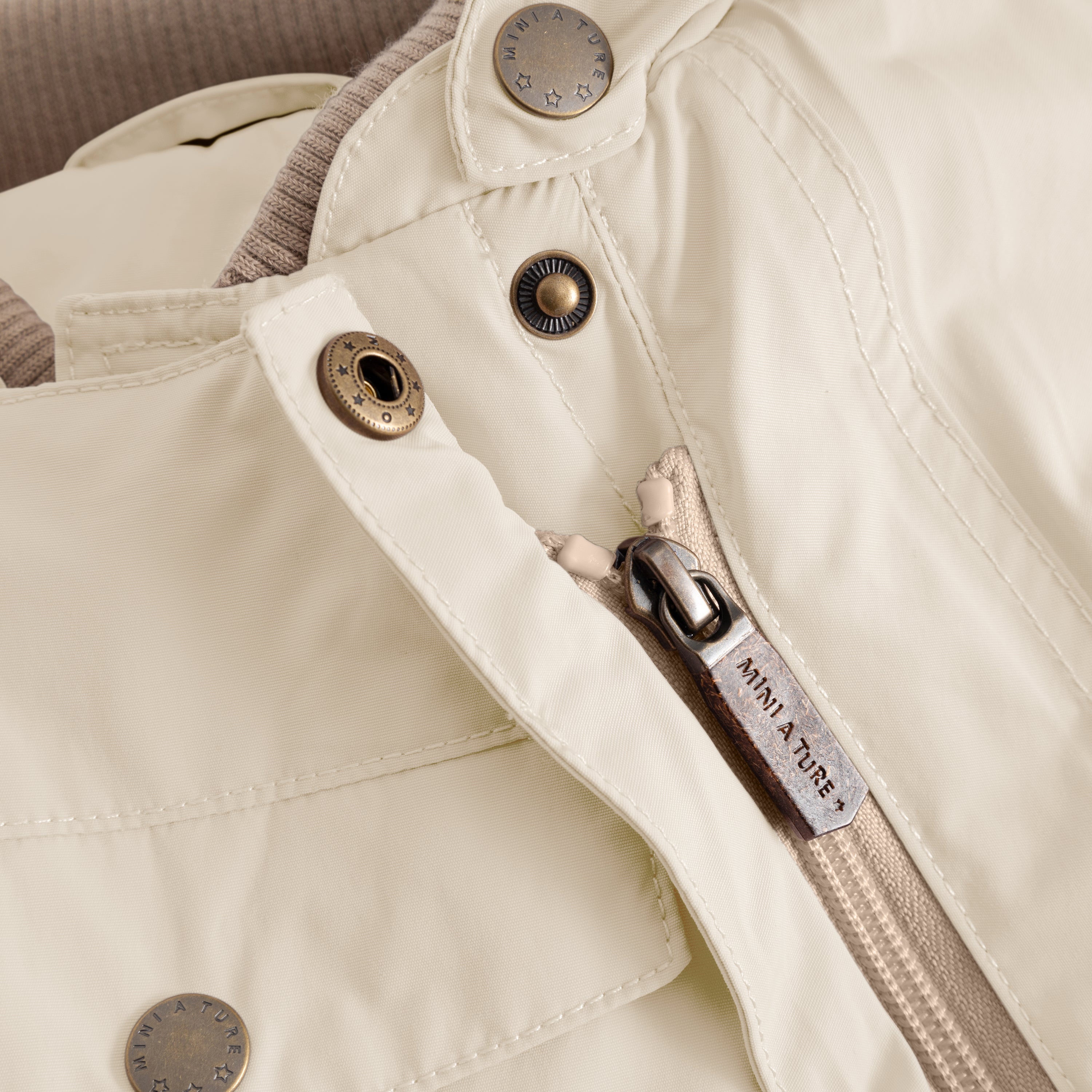 Close-up detail of the MATWISTI fleece lined snowsuit in warm beige showing one of the two slanted front zips with branded MINI A TURE metal zip pull, embossed metal snap buttons and beige ribbed cuff visible at the sleeve opening.