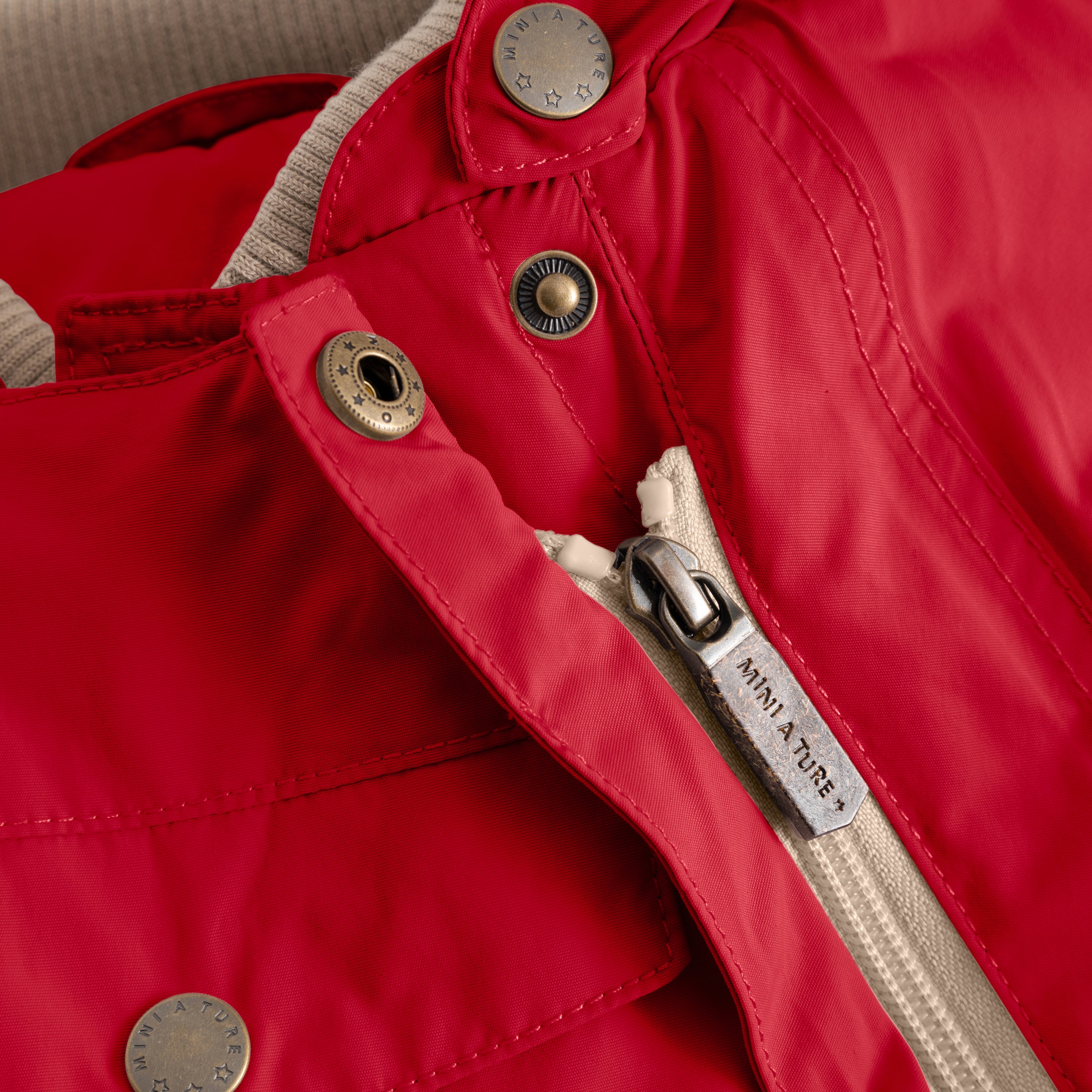 Close-up of the MATWISTI fleece lined snowsuit in bright red showing the slanted front zip with branded MINI A TURE metal zip pull, embossed metal snap buttons and beige fleece lining visible at the collar opening.
