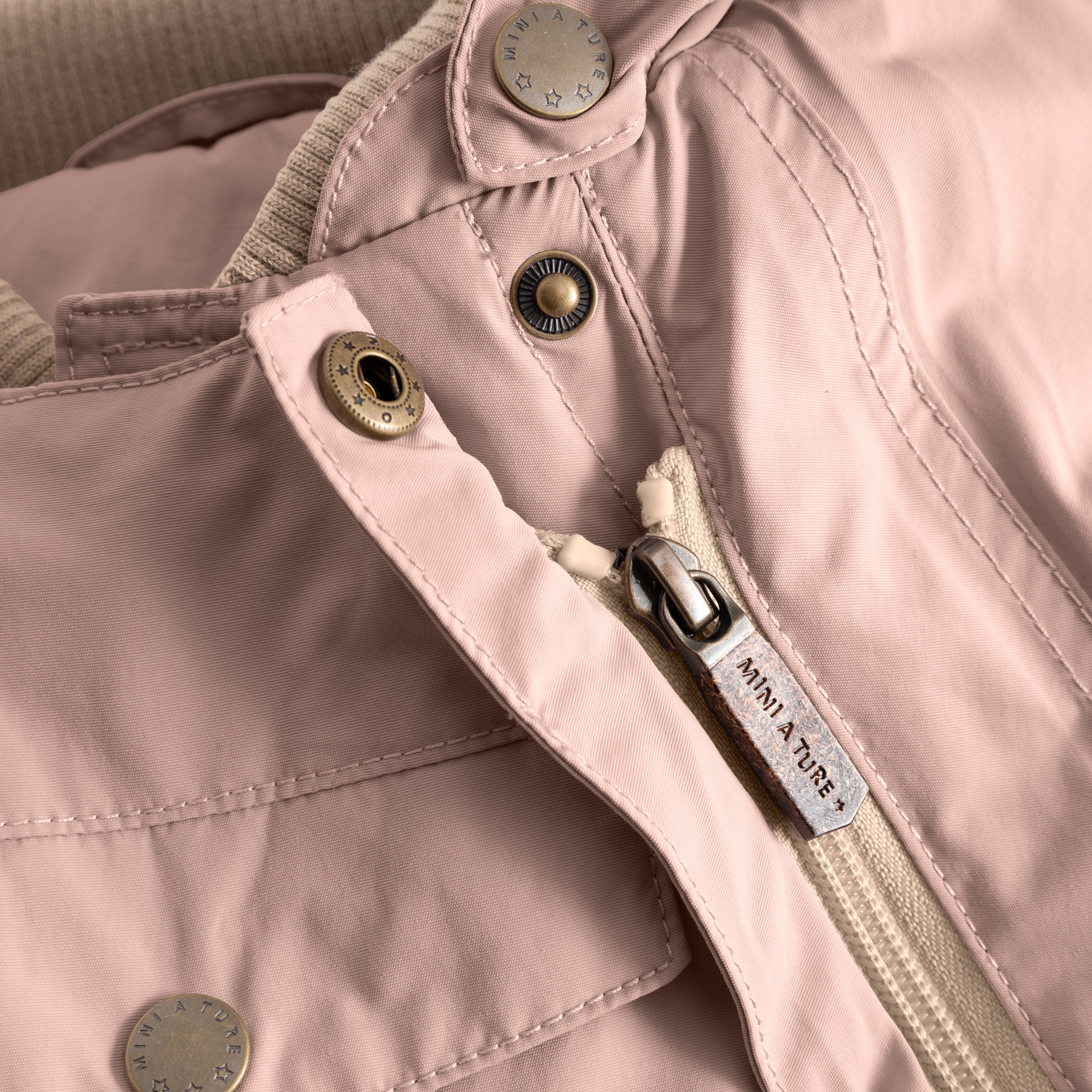 Close-up of the MATWISTI fleece lined snowsuit in soft rose pink showing the embossed MINI A TURE metal snap buttons, branded zip pull with beige fabric tab and front chest pocket with visible stitching detail.