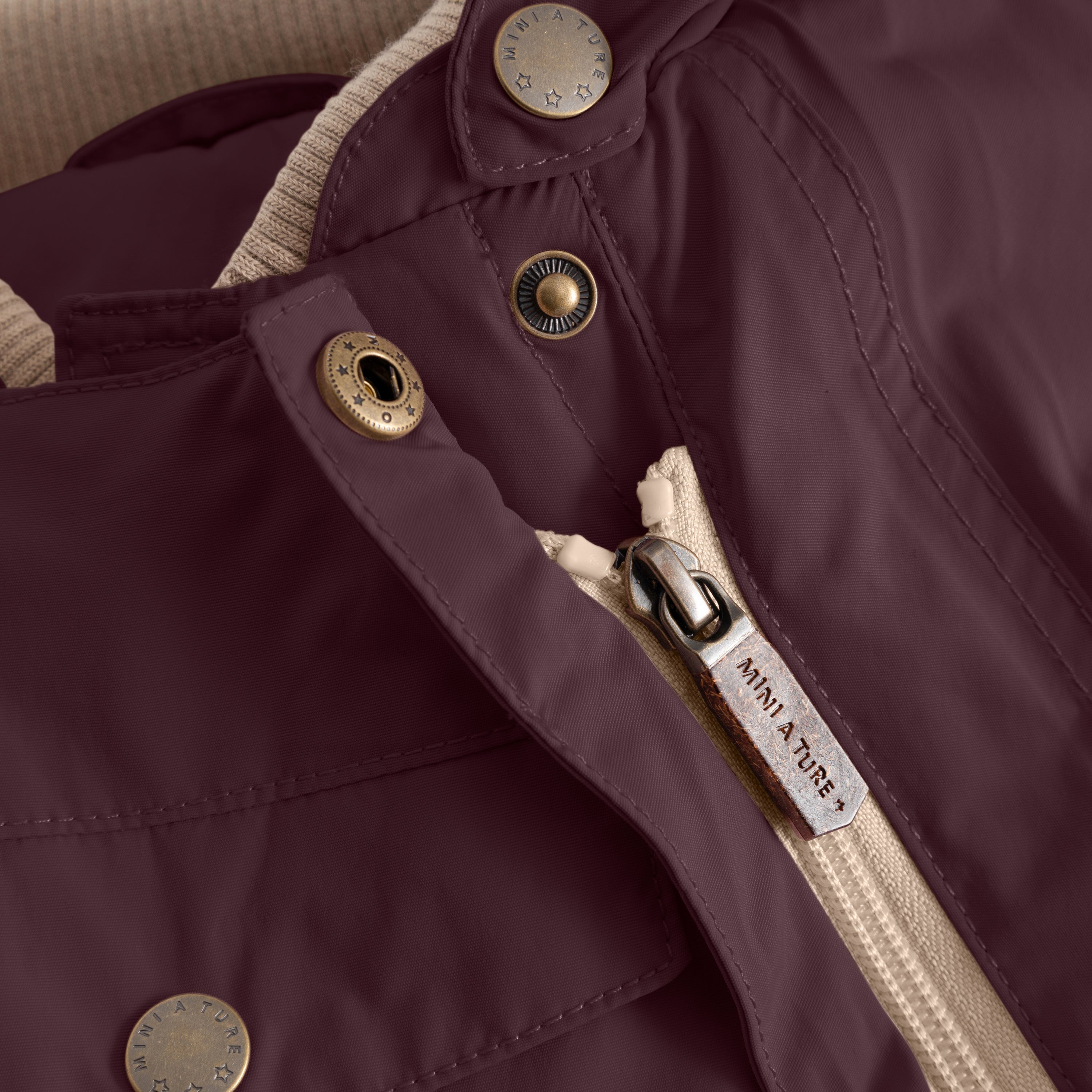 Close-up detail of the MATWISTI fleece lined snowsuit in deep plum showing one of the two slanted front zips with MINI A TURE branded zip pull, snap-button fastening and the brand's embossed metal stud at the collar.