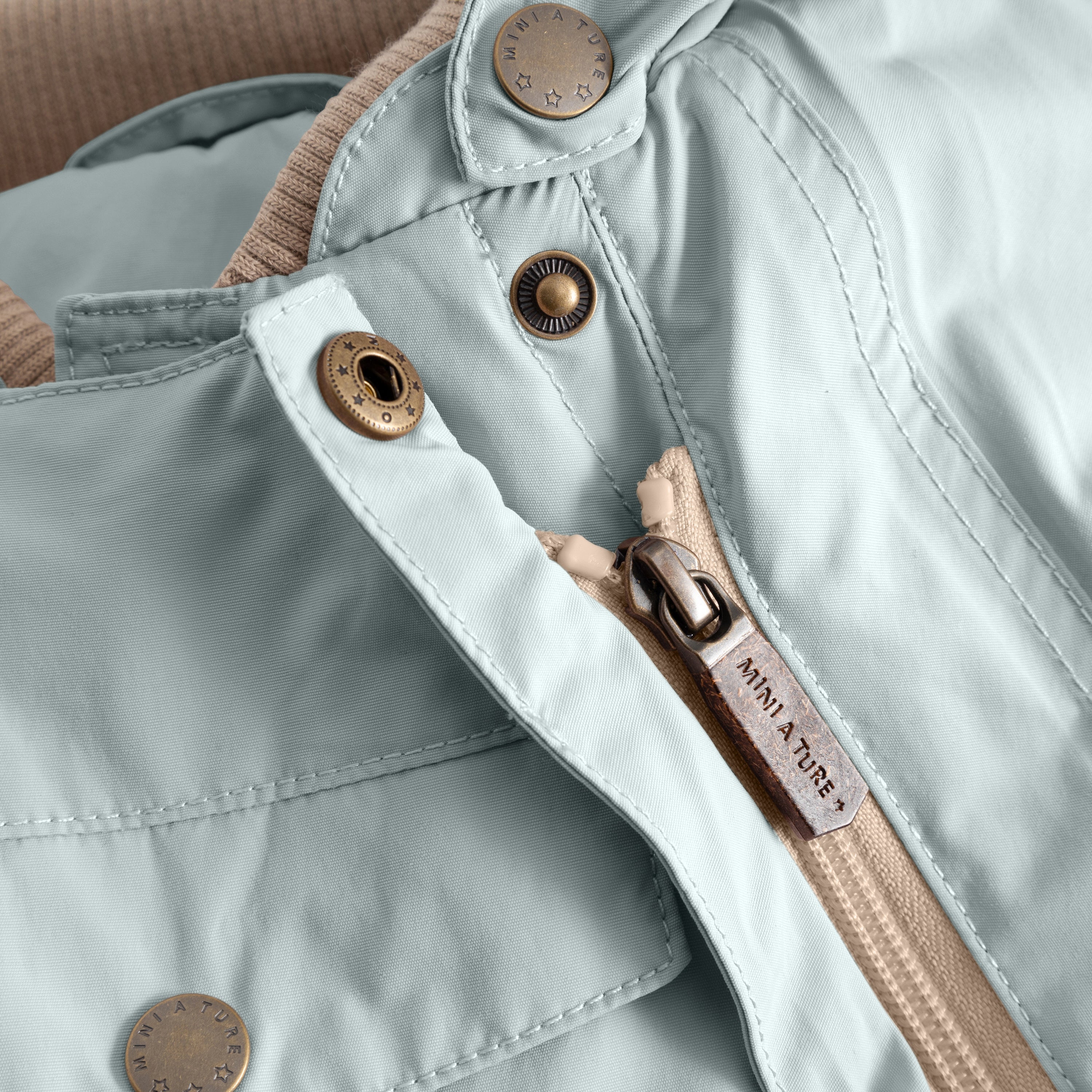 Close-up of the MATWISTI fleece lined snowsuit in pale blue showing the slanted front zip with branded MINI A TURE metal zip pull, embossed metal snap buttons and beige fleece lining visible at the collar opening.