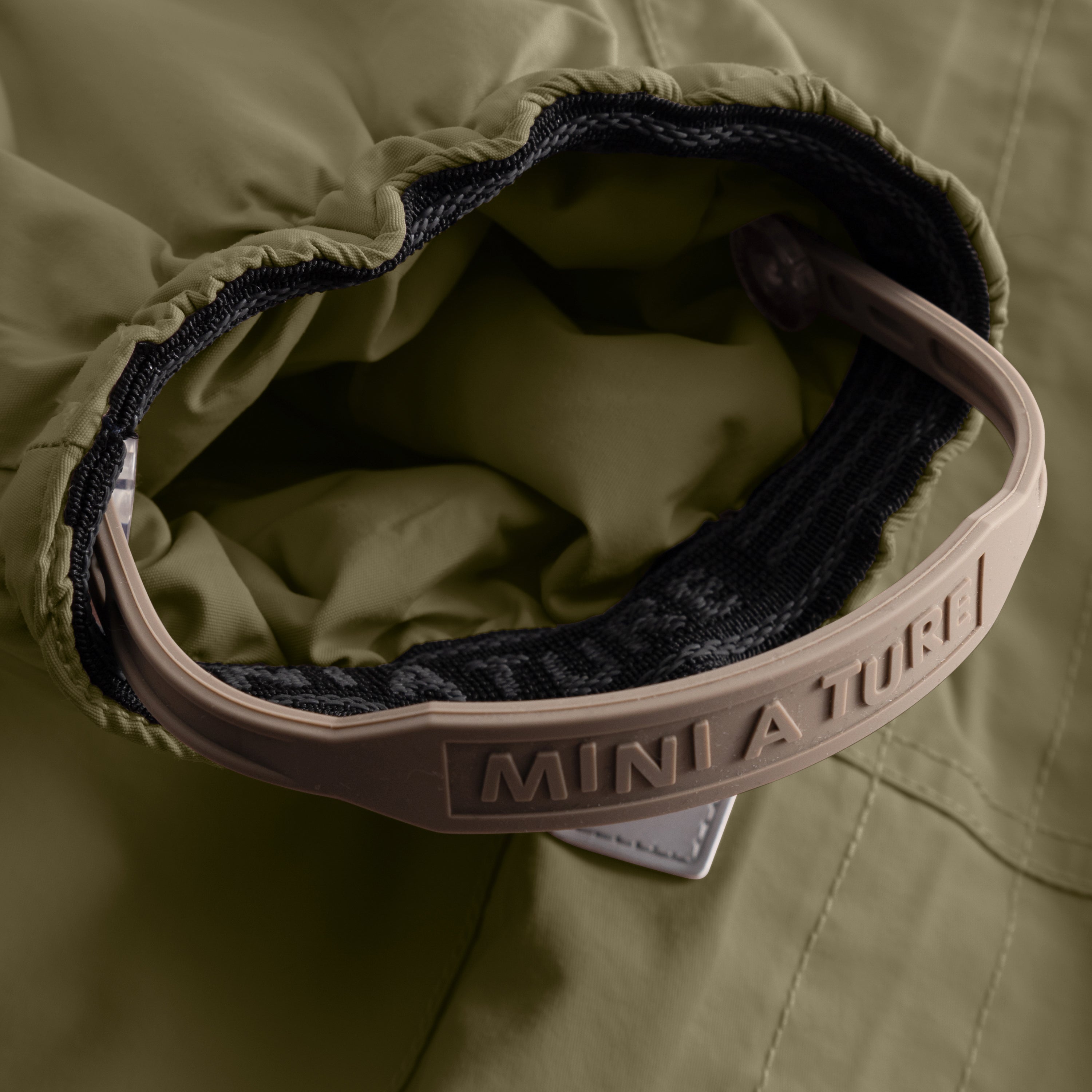 Close-up of the MATWISTI fleece lined snowsuit in olive green showing the adjustable foot strap with embossed MINI A TURE branding on the beige plastic buckle, black elasticated cuff and soft fleece lining visible inside the leg opening.