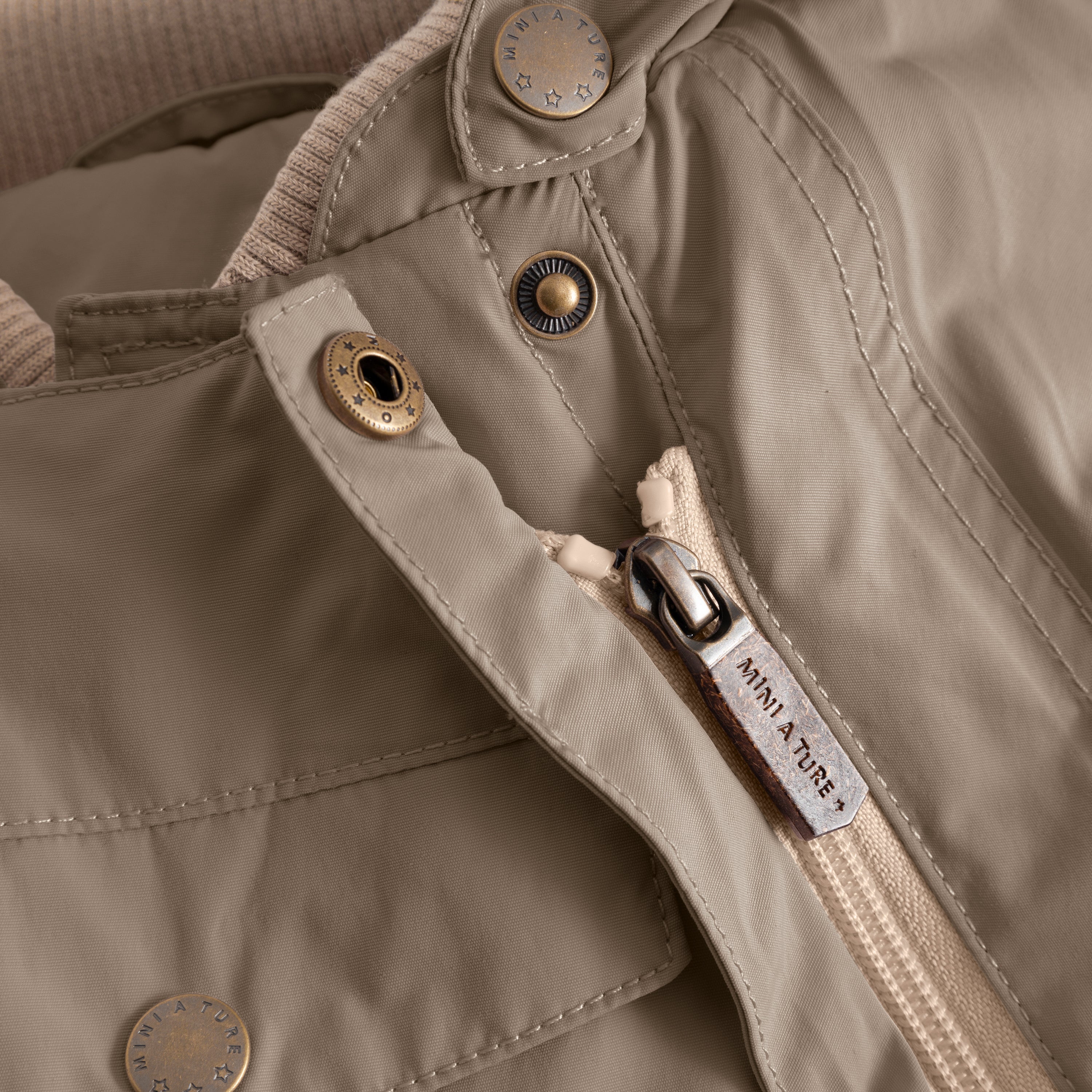 Close-up of the MATWISTI fleece lined snowsuit in warm beige showing the slanted front zip with branded MINI A TURE metal zip pull, embossed metal snap buttons and beige fleece lining visible at the collar opening.