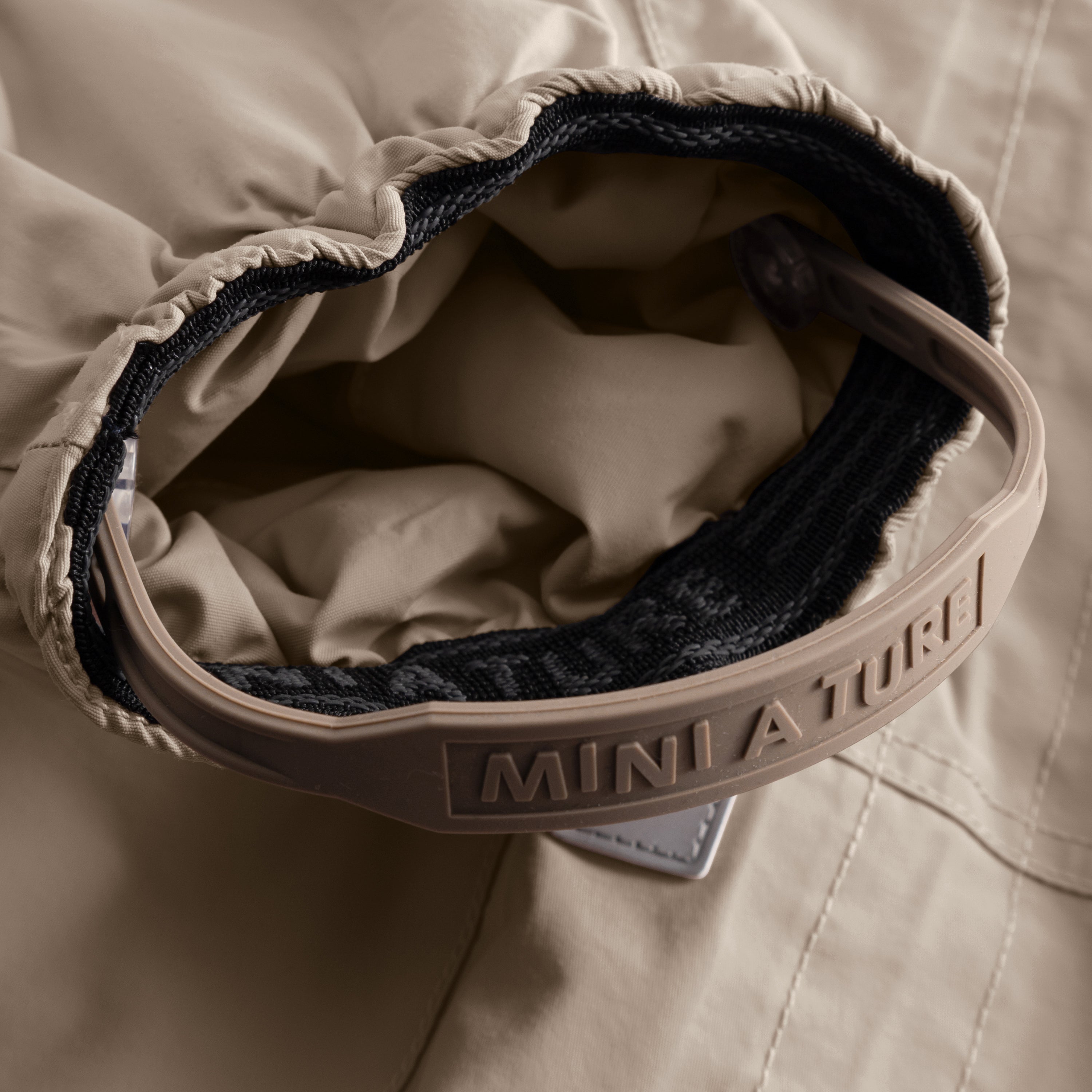 Close-up of the MATWISTI fleece lined snowsuit in warm beige showing the adjustable foot strap with embossed MINI A TURE branding on the beige plastic buckle, black elasticated cuff and soft fleece lining visible inside the leg opening.