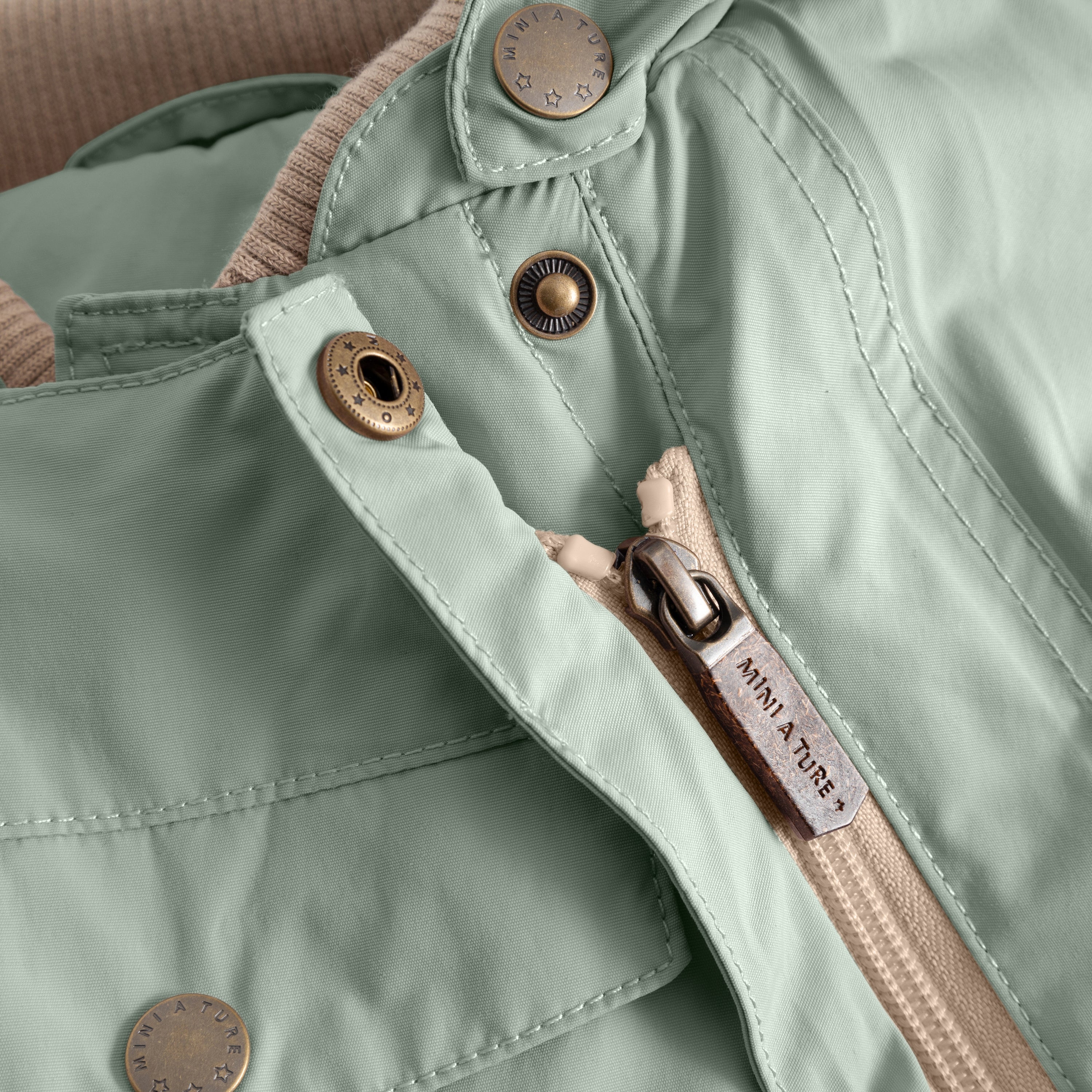 Close-up detail of the MATWISTI fleece lined snowsuit in sage green, showing one of the two hidden slanted front zips with branded metal snap buttons, adjustable foot straps and MINI A TURE label on the zip pull.