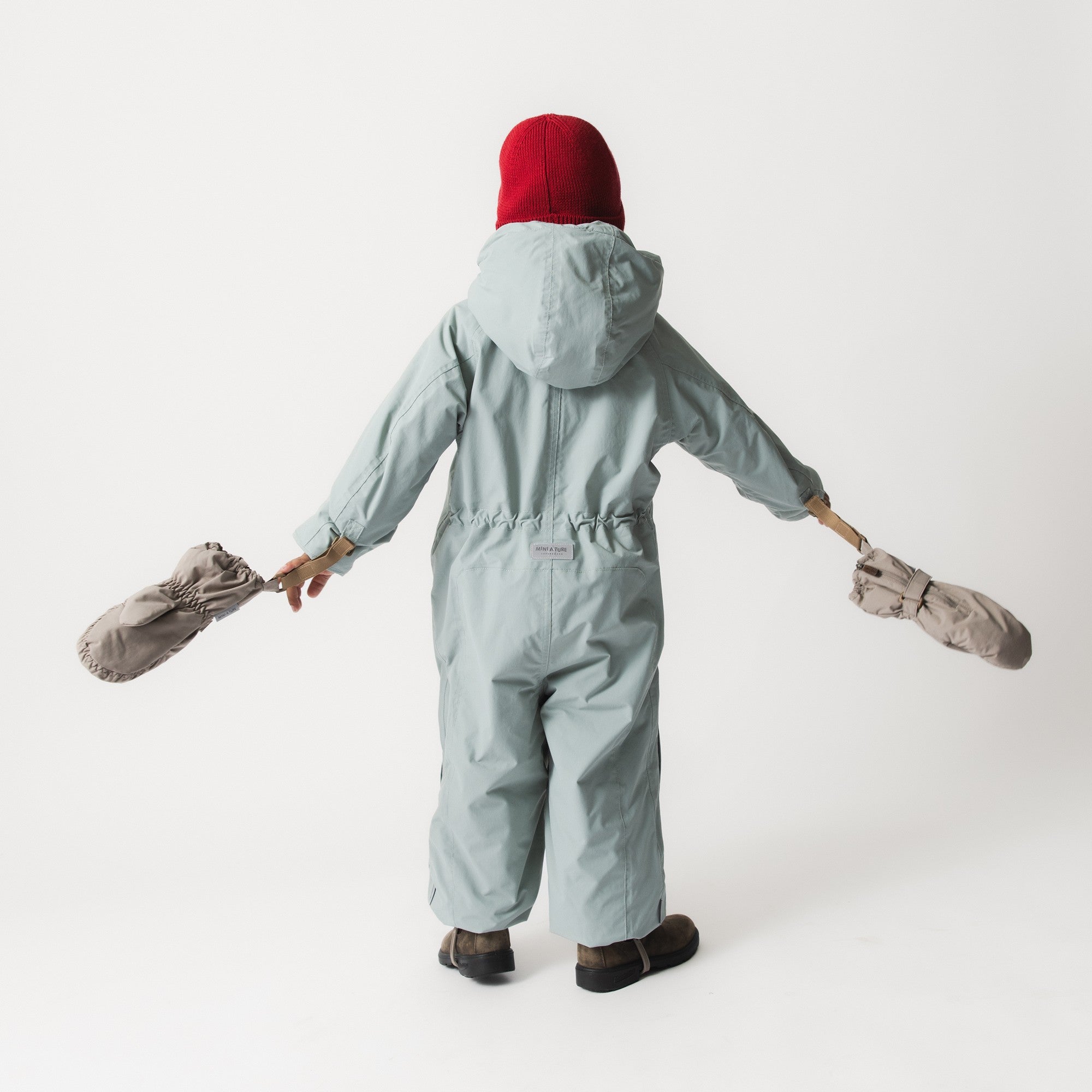 Rear view of a child wearing the MATWISTI fleece lined snowsuit in pale blue with a red knitted beanie, arms extended holding matching mittens, showing the detachable hood, elasticated waist and MINI A TURE label at the lower back.