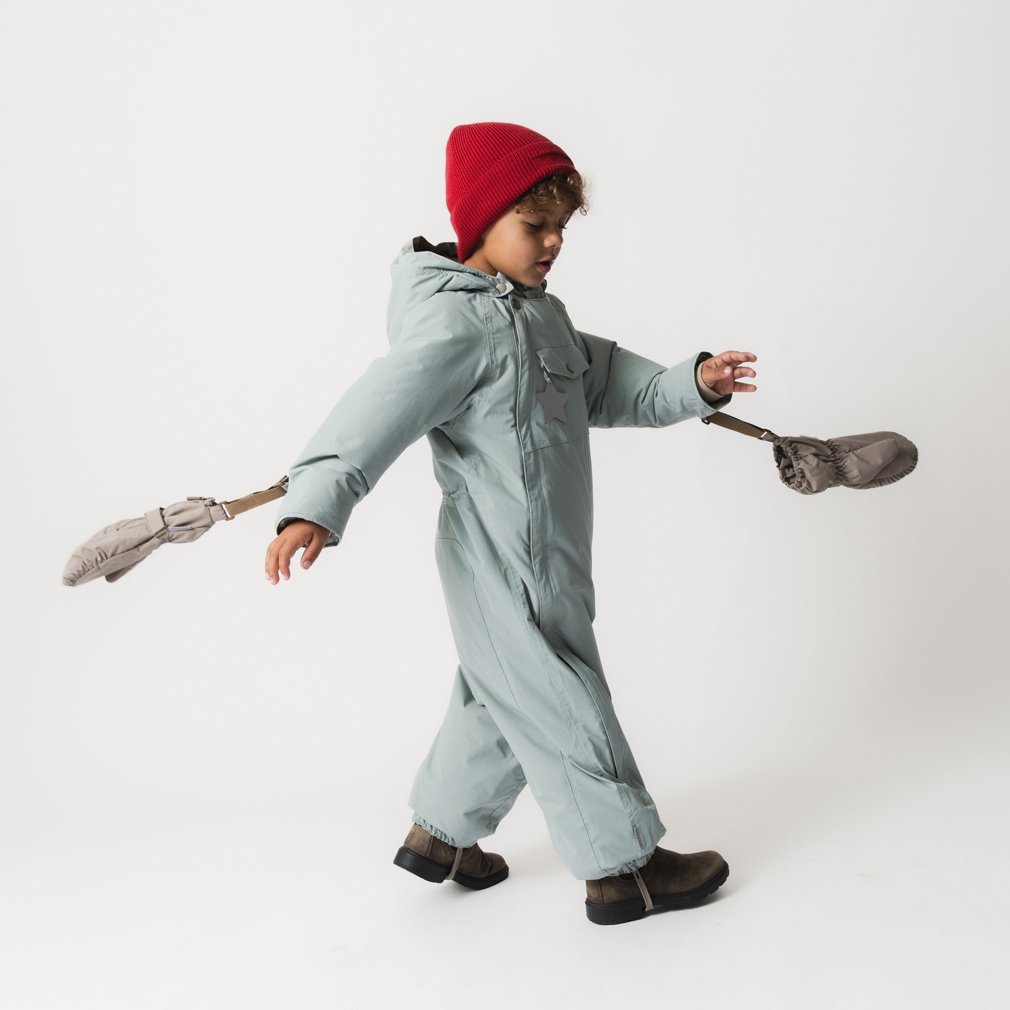 Child in motion wearing the MATWISTI fleece lined snowsuit in pale blue with arms outstretched, paired with a red knitted beanie and brown boots, holding mittens by their straps.