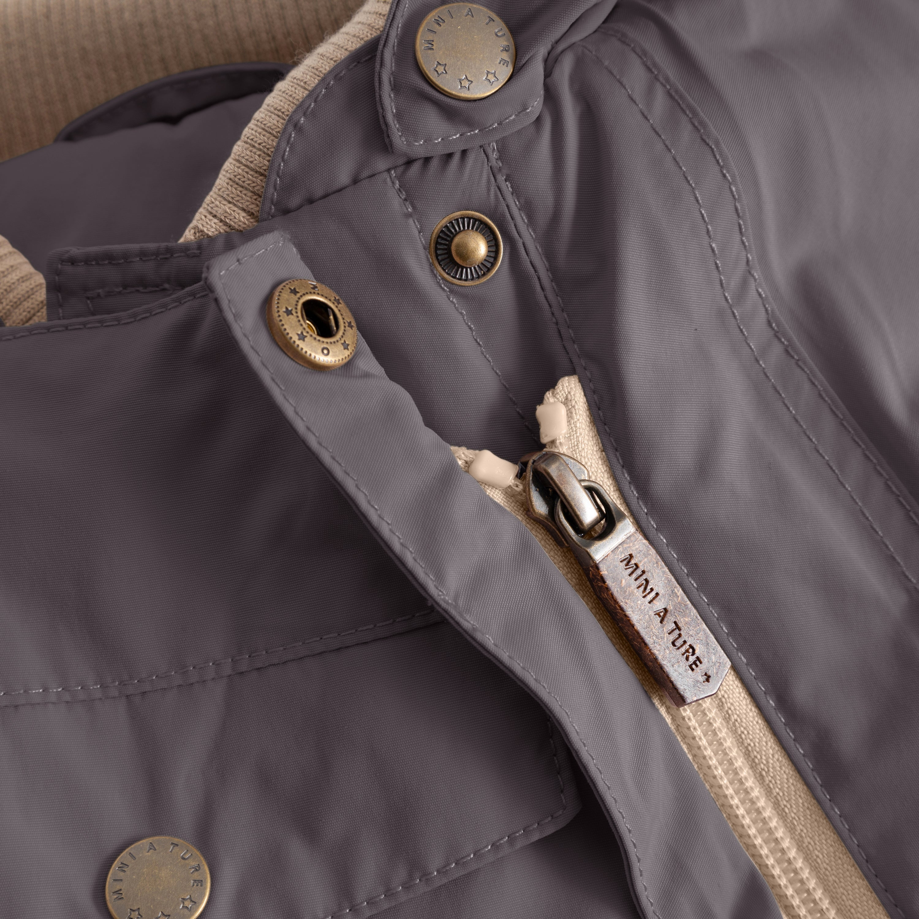 Close-up of the MATWISTI fleece lined snowsuit in charcoal grey showing the embossed MINI A TURE metal snap buttons, branded zip pull with beige fabric tape and front chest pocket with visible stitching detail.