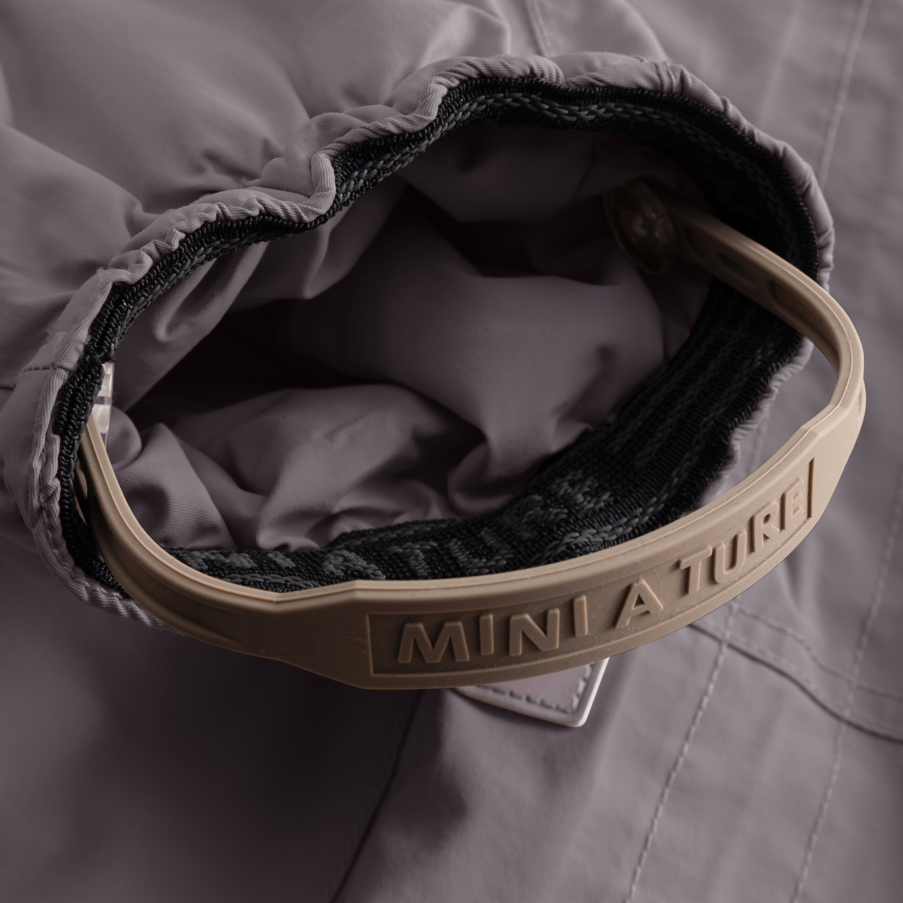 Close-up of the MATWISTI fleece lined snowsuit in charcoal grey showing the adjustable foot strap with embossed MINI A TURE branding on the beige plastic buckle, black elasticated cuff and soft fleece lining visible inside the leg opening.