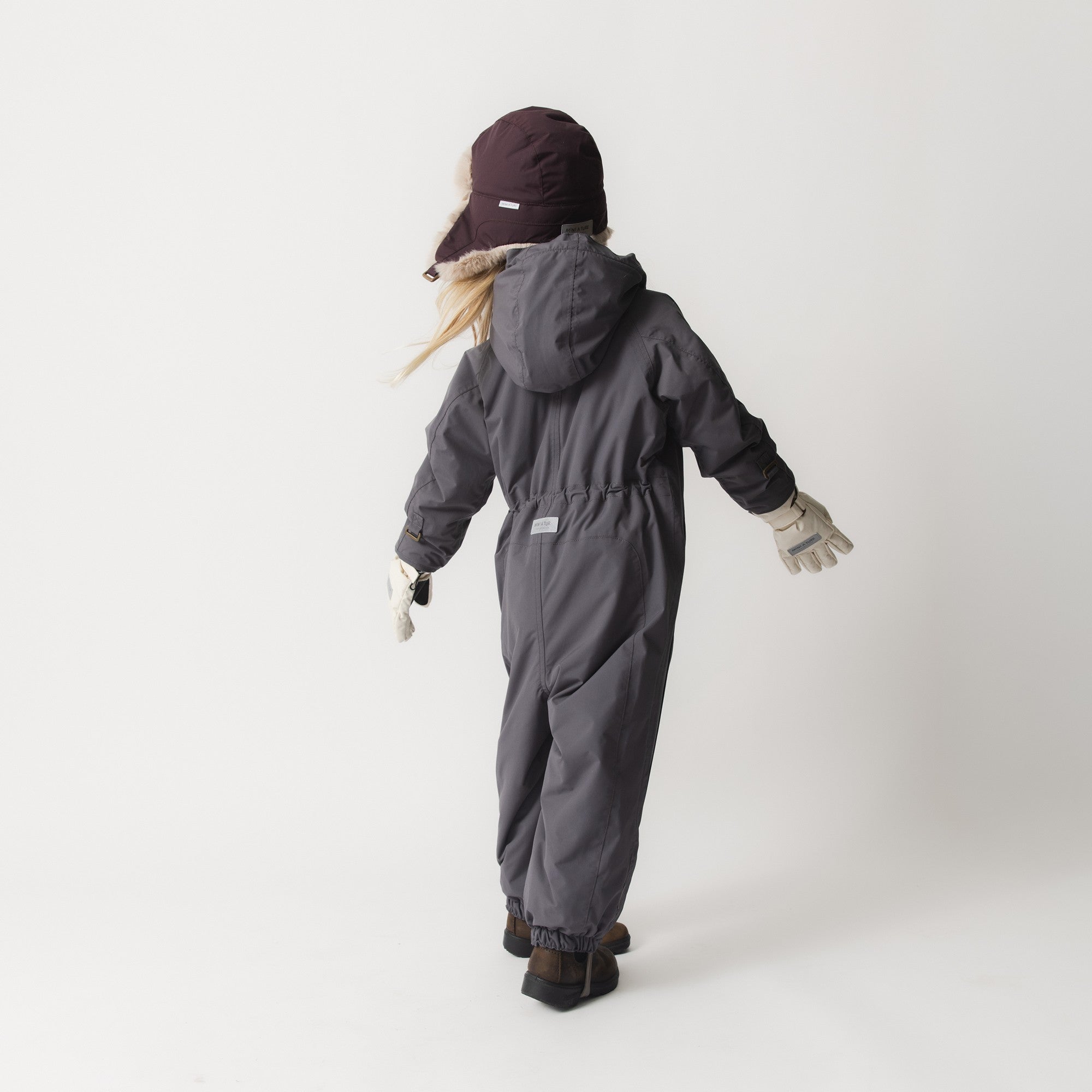 Young child wearing the MATWISTI fleece lined snowsuit in charcoal grey from behind, showing the detachable hood with faux-fur trim, internal waist adjustment, beige ribbed cuffs and elasticated leg openings with adjustable foot straps during active movement.