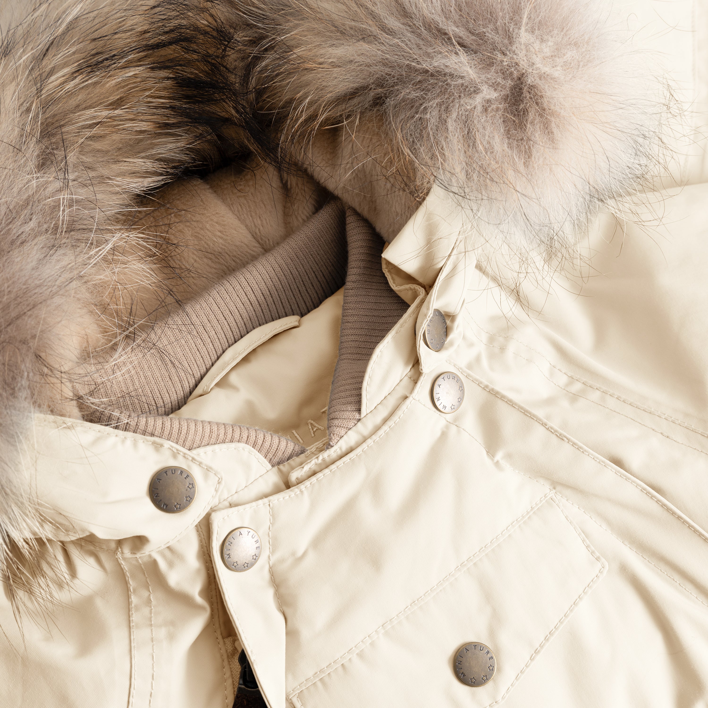 Close-up of MATWISTI fleece lined snowsuit fur in cream showing the detachable genuine fur hood trim, taupe ribbed collar, MINI A TURE branded press-stud fastenings along the chest, and front pocket detail on the waterproof shell.