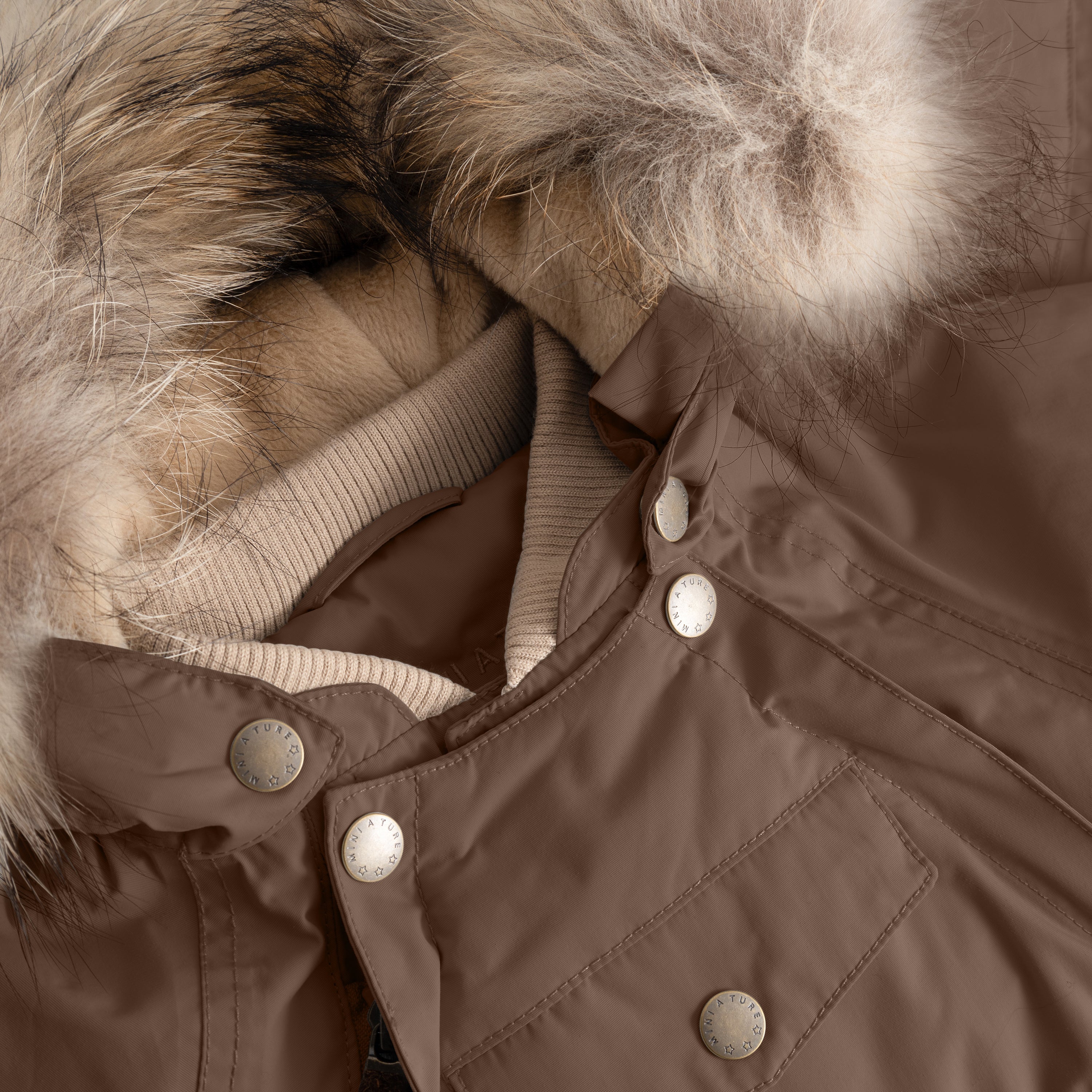 Close-up of MATWISTI fleece lined snowsuit fur in brown showing the genuine fur hood trim, beige ribbed collar, MINI A TURE branded press-stud fastenings along the chest, and front pocket detail on the waterproof shell.